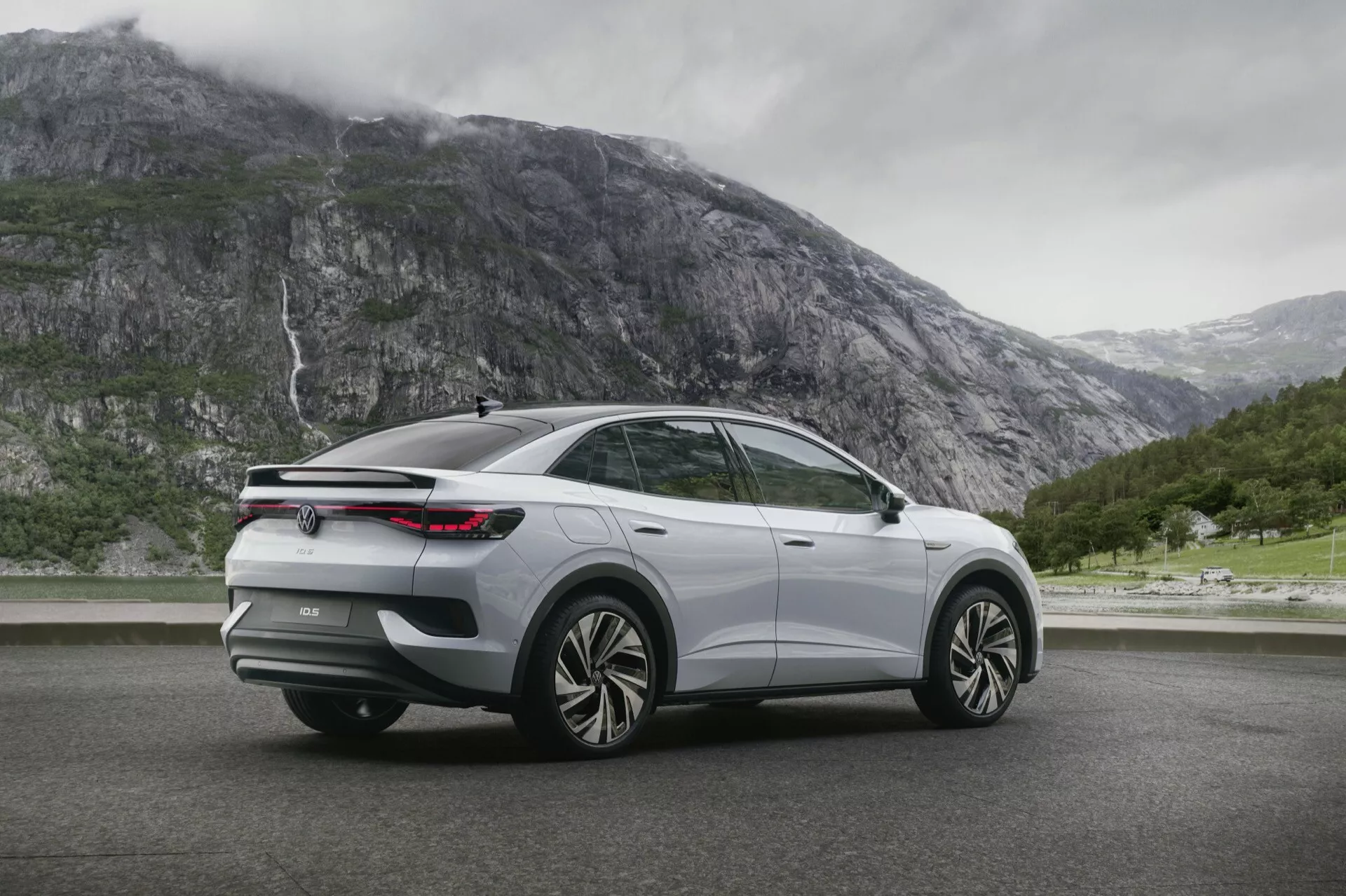 VW ID5 SUV 2022 zachycený v moderním třičtvrtinovém pohledu zezadu. Obrázek zdůrazňuje pravý bok vozidla z vyvýšeného úhlu a ukazuje velká kola z lehkých slitin s víceramenným designem, elegantní zadní LED světla, klesající linii střechy se zadním spoilerem, zatmavená okna, stříbrný nebo světle šedý lak s chromem nebo leskle černým obložením. Vozidlo je umístěno na suché silnici s horami a vodopádem v pozadí, což naznačuje dobrodružný a venkovní orientovaný nádech.
