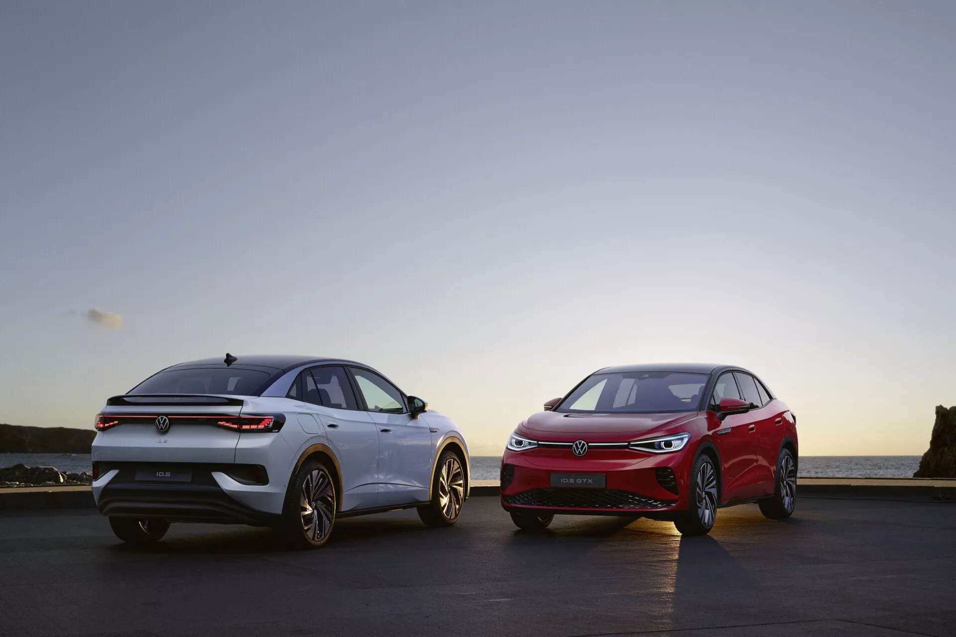 Obraz ukazuje dvě auta zaparkovaná vedle sebe. Auto vlevo, pravděpodobně VW ID.5 2022 (1. generace), je zobrazeno z tříčtvrtečního pohledu zezadu, zvýrazňující jeho záď, zadní kolo, část strany spolujezdce a pohled na předek zezadu. Vypadá, že je světle zbarvené s výraznými zadními světly, zadním oknem, nárazníkem a pravděpodobně zadním difuzorem. Auto vpravo, tmavší barvy, je prezentováno z tříčtvrtečního pohledu zepředu, zvýrazňující jeho přední mřížku, světlomety, stranu řidiče a část předního kola. Mřížka může mít senzory pro pokročilé asistenty řidiče. Scéna je zasazena na rovném povrchu u vodního plochy během časného rána nebo pozdního večera.