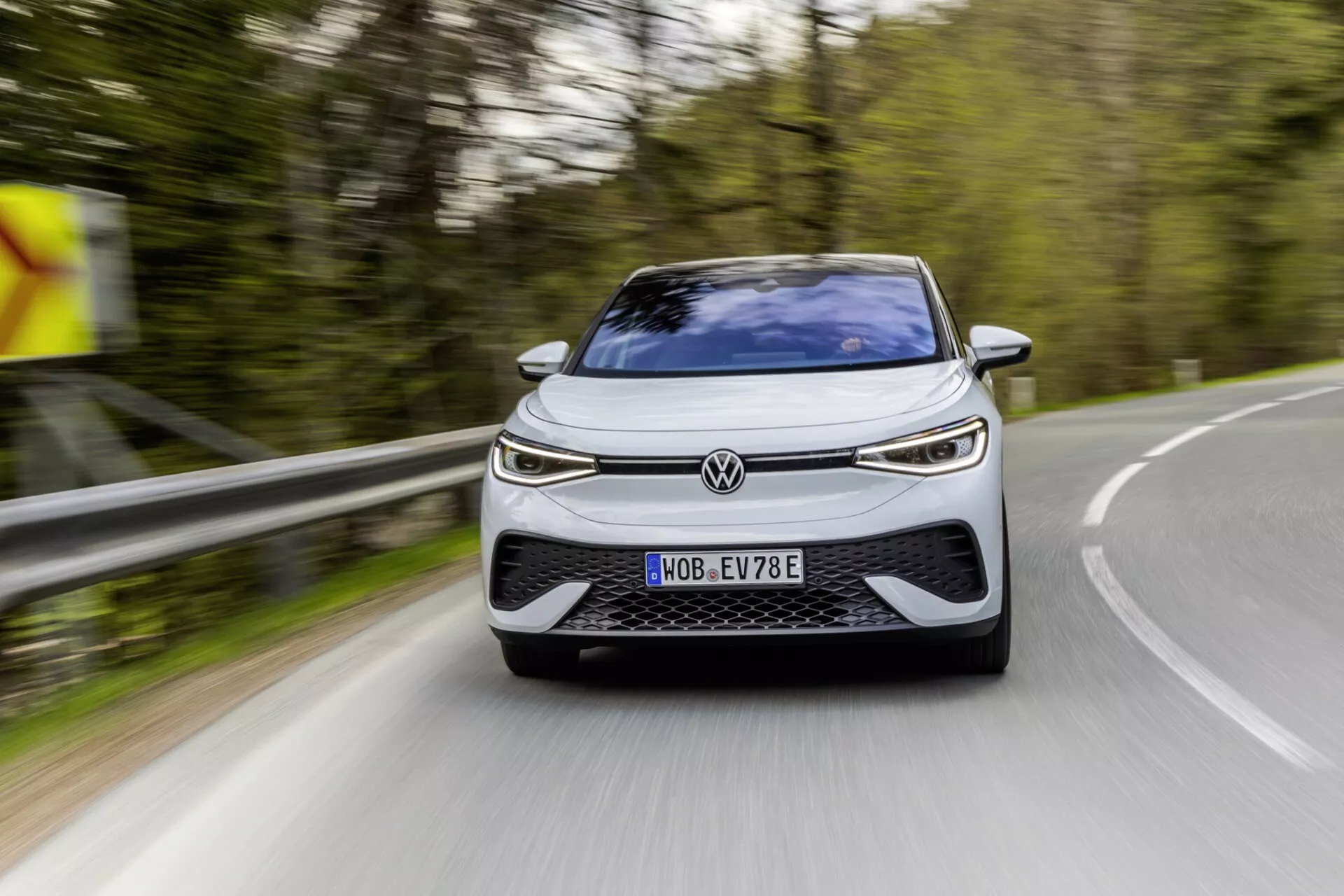 Volkswagen ID.5 2022 (1. generace) zachycený na venkovské silnici, ukazující nízký přední úhel se zaměřením na elegantní přední část zahrnující mřížku, LED světlomety a aerodynamický nárazník. Auto se pohybuje mírně doprava a vpřed, což ukazuje jasné designové a technologické prvky naznačující elektrické nebo hybridní vozidlo, registrované v Německu, s moderním dynamickým vzhledem.