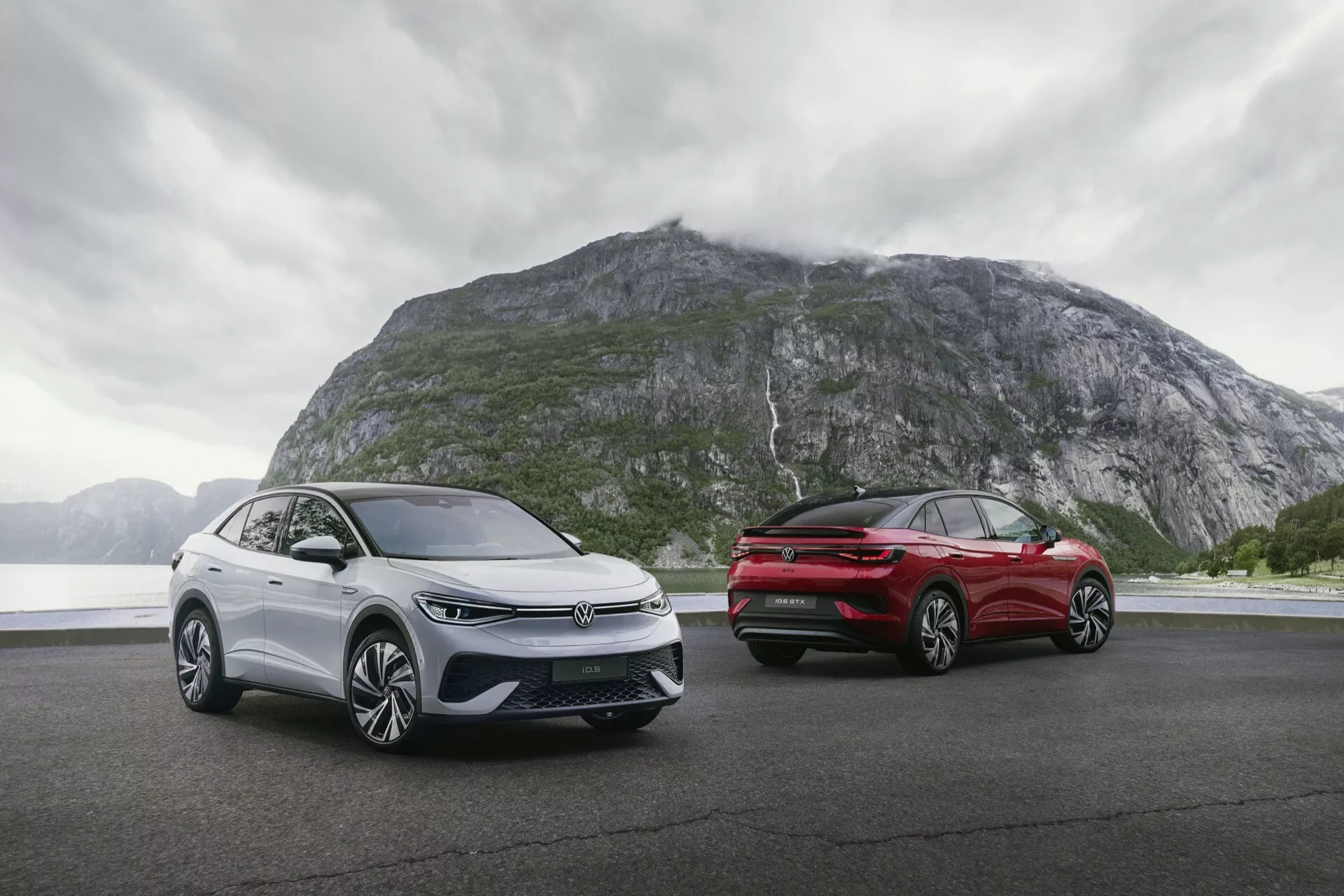 Dva vozy VW ID.5 2022 (1. generace) zaparkované na rovném povrchu s dramatickým horským pozadím. Pochmurné počasí s dešťovými mraky přidává na náladě. Vůz vlevo je zobrazen z tři čtvrtinového předního úhlu s úzkými LED světlomety, uzavřenou mřížkou, senzory ADAS a aerodynamickými koly. Vůz vpravo je vidět z tři čtvrtinového zadního úhlu s úzkými LED zadními světly, zadním difuzorem a elegantním designem. Oba vozy mají litá kola, metalický povrch a elegantní boční zrcátka s integrovanými směrovkami.