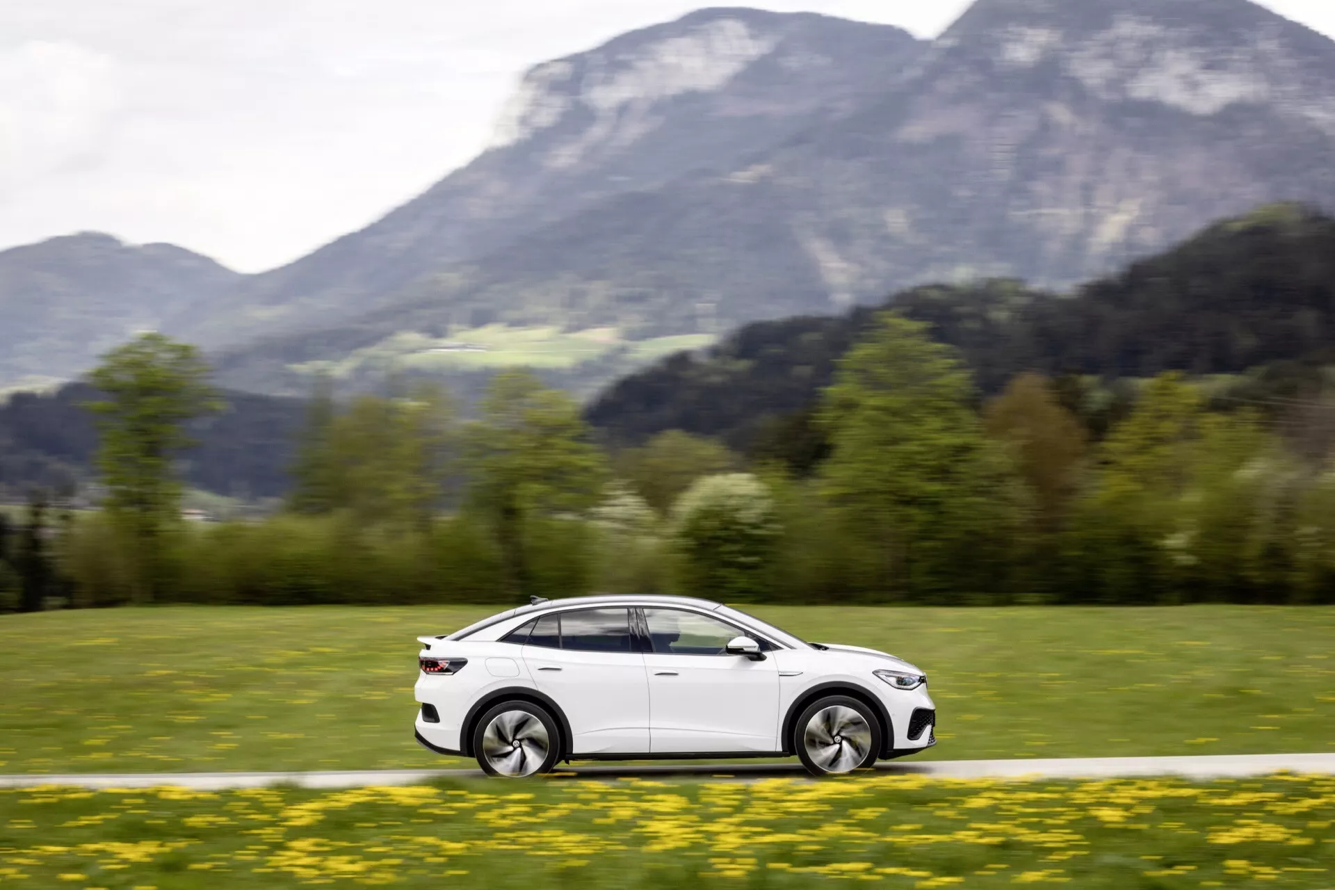 Obrázek zobrazuje dynamický boční profil VW ID5 2022 (1. generace) v pohybu, zdůrazněný rozmazáním kol a pozadí. Fotografie z mírného nadhledu a střední vzdálenosti zvýrazňuje hladký a aerodynamický design vozu, včetně dveří, bočních oken, zrcátek a designových linií. Kola jsou v pohybu a mírně natočená, což naznačuje zatáčení nebo manévr. Odrážení světla ukazuje na lesklý, dobře udržovaný exteriér. Vůz je zasazen do malebné venkovní krajiny s trávící zelení v popředí a horami v pozadí, což zvyšuje pocit rychlosti a dobrodružství.