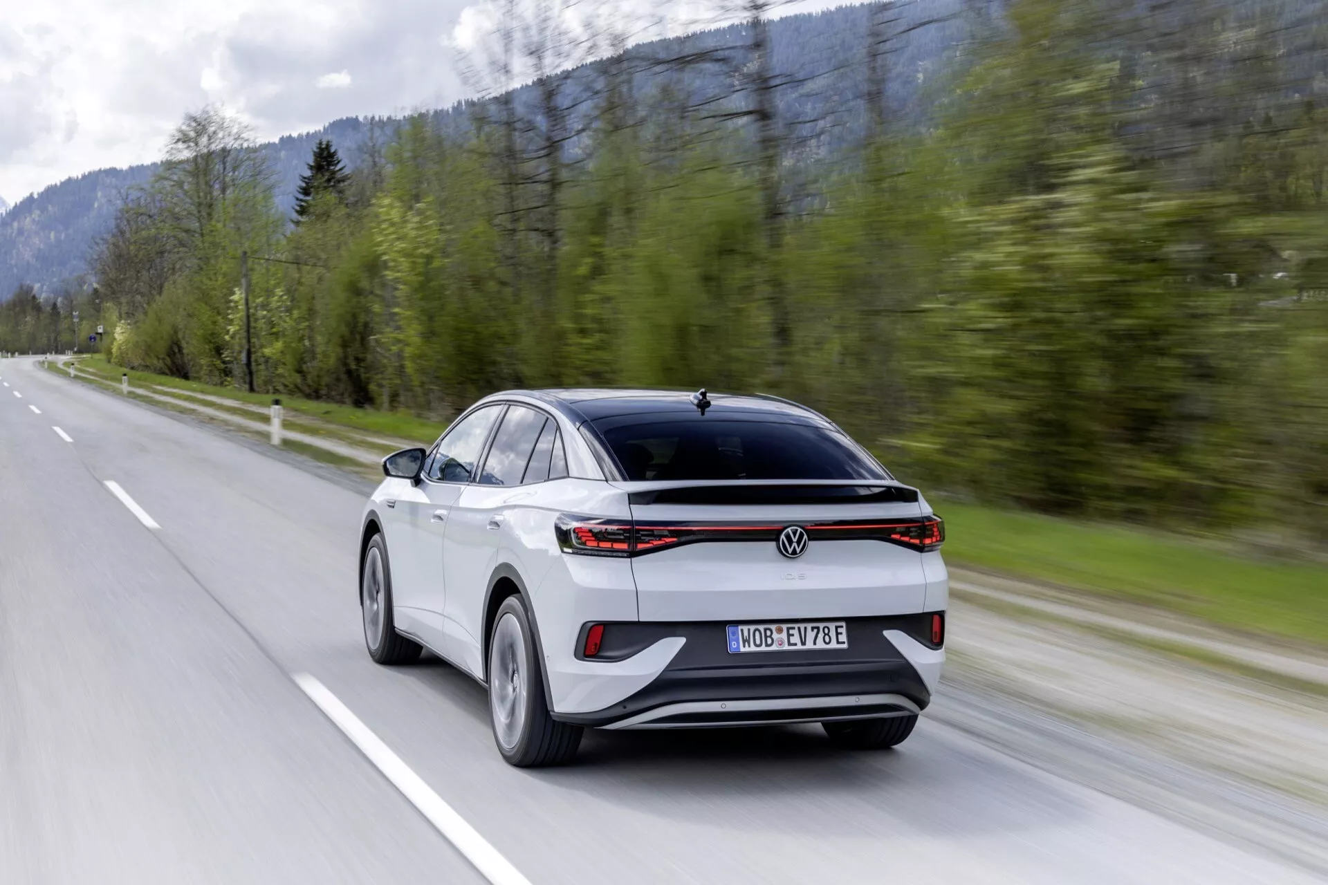 VW ID.5 2022 první generace moderní crossover/SUV, zachycený z tříčtvrtečního zadního pohledu ukazující sportovní a aerodynamický design se skloněným zadním oknem, charakteristickým LED zadním světelným pruhem, dvoubarevným bílým a černým kontrastním vzhledem, prémiovými multifunkčními slitinovými koly, senzory pokročilého asistenčního systému řidiče, centrální registrační značkou a logem, zachycen v pohybu na dvouproudé venkovské silnici s lesnatým horským pozadím, které evokuje rychlost a výkon, pravděpodobně elektrické nebo hybridní vozidlo.