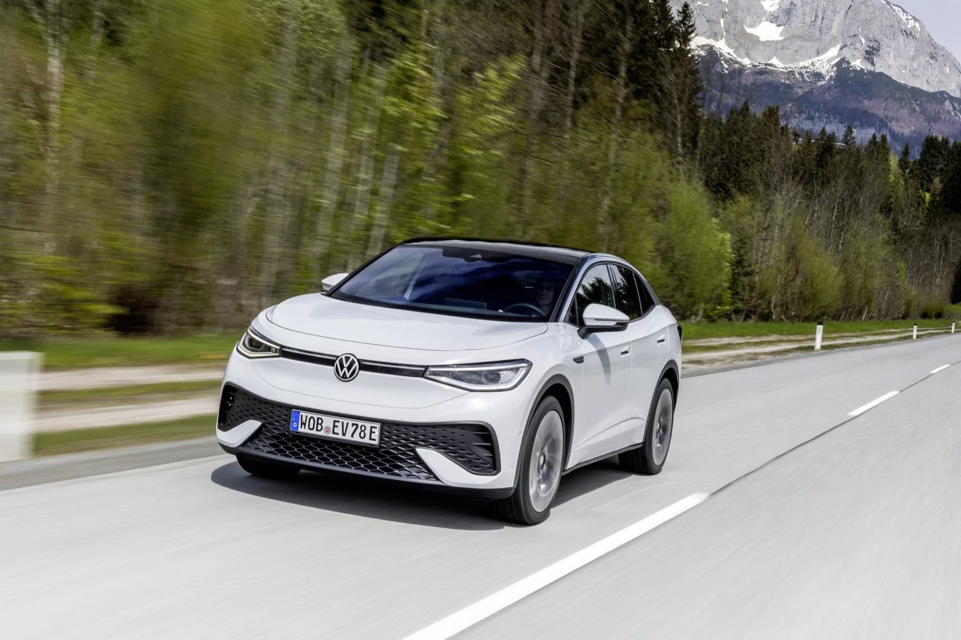 Obrázek moderního VW ID.5 2022 (1. generace) elektrického vozidla, zachyceného za denního světla v pohybu, zobrazující pohled z předního a bočního profilu pod úhlem 45 stupňů. Auto, hatchback nebo kompaktní SUV, je zvýrazněno svým výrazným předním grillem a úzkými horizontálními světlomety. Vozidlo vykazuje funkce jako denní svícení, aerodynamické ráfky, boční zrcátka s integrovanými směrovými světly, panoramatické střešní okno a dvoubarevný lak. Rozmazané pozadí a povrch silnice ve fotografii zvýrazňují pohyb vozu, zatímco 'E' na registrační značce jej identifikuje jako elektrický model.