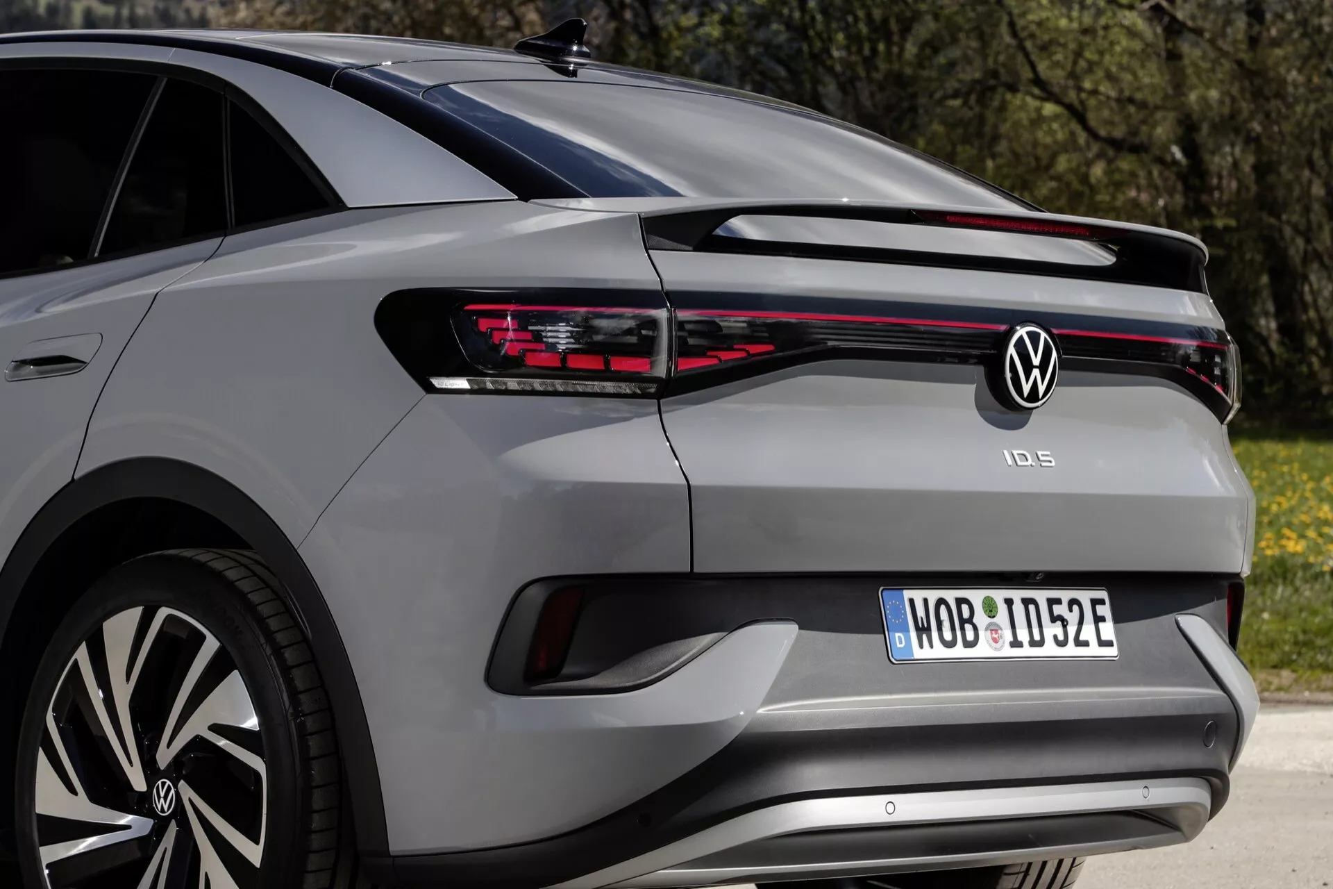 Zadní třičtvrteční úhel pohledu na moderní hatchback VW ID.5 2022 (1. generace) ukazující LED zadní světlo, které se táhne po celé šířce, výrazné segmentované zadní světla na obou stranách, centrální znak značky, název modelu pod světlem, registrační značku v jejím výklenku, zadní nárazník s parkovacími senzory, stylový zadní spoiler s namontovaným stíračem a část vícepaprskového hliníkového kola, odrážející exteriérové designové prvky a technologie.