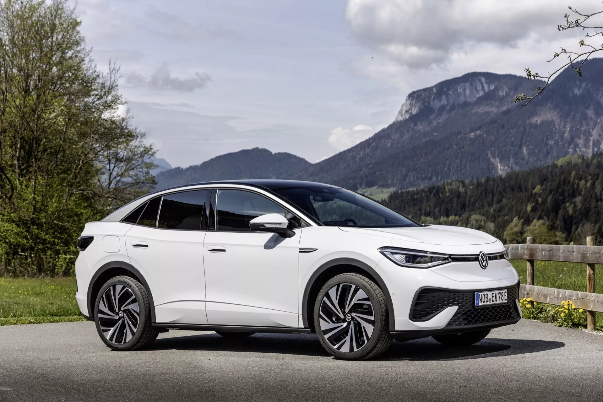Moderní bílá SUV VW ID.5 z roku 2022 zaparkovaná venku v malebném prostředí se zelení a horami. Vozidlo je zachyceno z tříčtvrtečního předního pohledu a zvýrazňuje přední mřížku, LED světlomety, slitinová kola a šikmou linii střechy. Obraz naznačuje pokročilé technologické funkce, jako jsou kamery namísto bočních zrcátek, a prémiový, aerodynamický design. Malebné prostředí podporuje vhodnost vozu pro městské i venkovské prostředí.