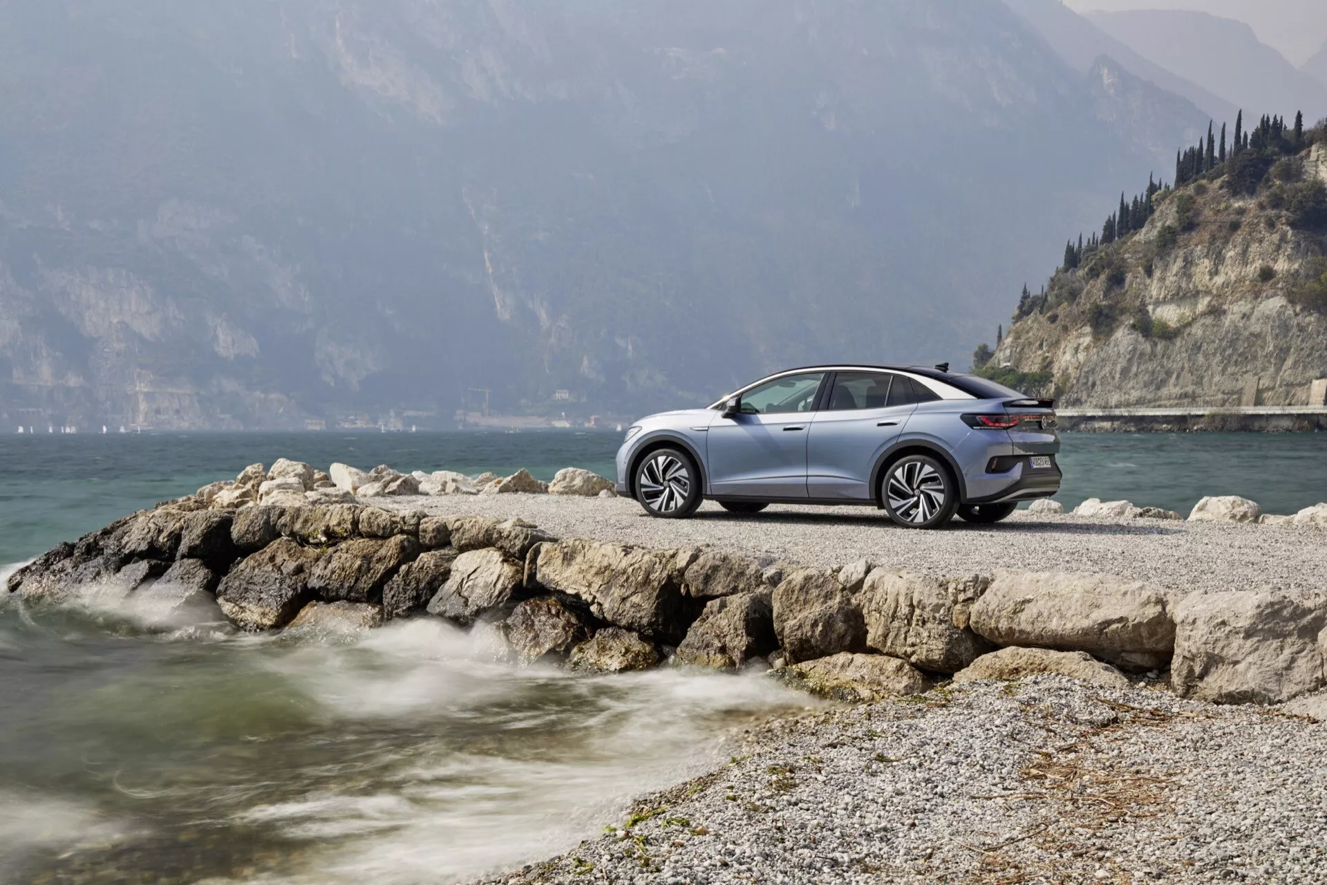 VW ID5 2022 (1. generace) auto zaparkované na skalnatém výběžku u vodní plochy s horským pozadím částečně zakrytým mlhou. Boční pohled na řidičovu stranu s vysokým podvozkem, velkými koly, LED světly, elegantními liniemi karoserie a metalickým lakem. Obrázek vyvolává pocit luxusu, moderního designu a dobrodružství v poklidném přírodním prostředí.
