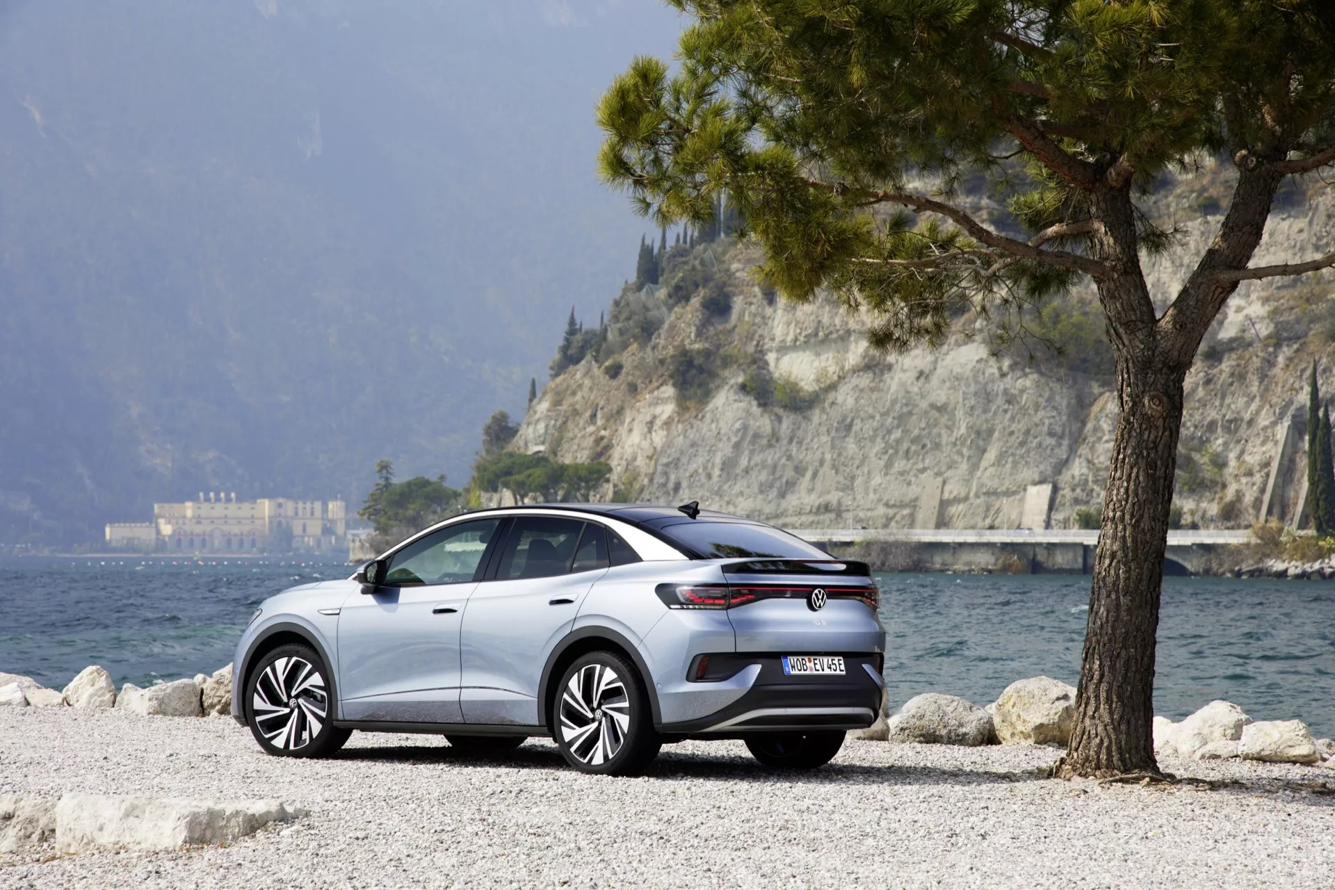 Moderní VW ID.5 2022, první generace, zaparkovaný na malebném venkovním místě u jezera. Pohled ze tří čtvrtin zezadu zvýrazňuje zadní a boční profil vozu, se zdůrazněním vícepaprskových litých kol, zadních světel, zpětných zrcátek, klik dveří a elegantních čar. Štěrkový povrch a strom naznačují jeho užitkový nebo crossover/SUV charakter. Dobře osvětlený za denního světla, snímek naznačuje pokročilé technologie vozidla, jako jsou asistenti řízení a funkce konektivity.