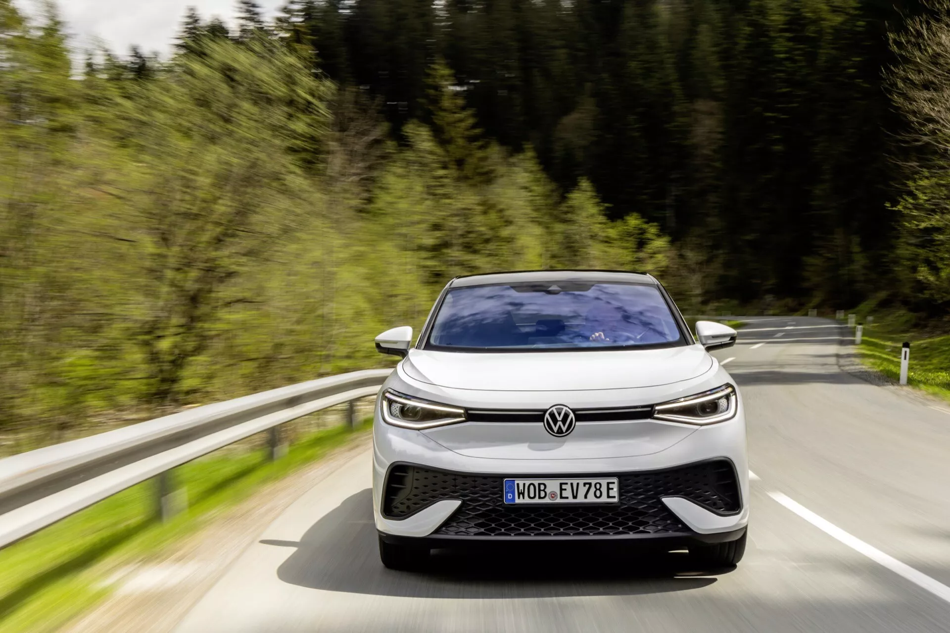 Obrázek zobrazuje Volkswagen ID.5 2022 (1. generace) z předního, mírně posunutého úhlu, jak se blíží k fotoaparátu. Moderní design auta zahrnuje nízký, široký postoj s výrazně zakřivenou kapotou, tenkou horizontální mřížkou, LED světlomety a velkými nasávacími otvory v předním nárazníku, což naznačuje výkonnostně orientovaný nebo agresivní design. Viditelná SPZ naznačuje, že automobil je na veřejné komunikaci, a rozmazání pohybu kolem vozidla a pozadí naznačuje, že auto je v pohybu v přírodním, snad lesnatém prostředí během dne. Písmeno 'E' na SPZ naznačuje, že se jedná o elektrické nebo hybridní vozidlo.