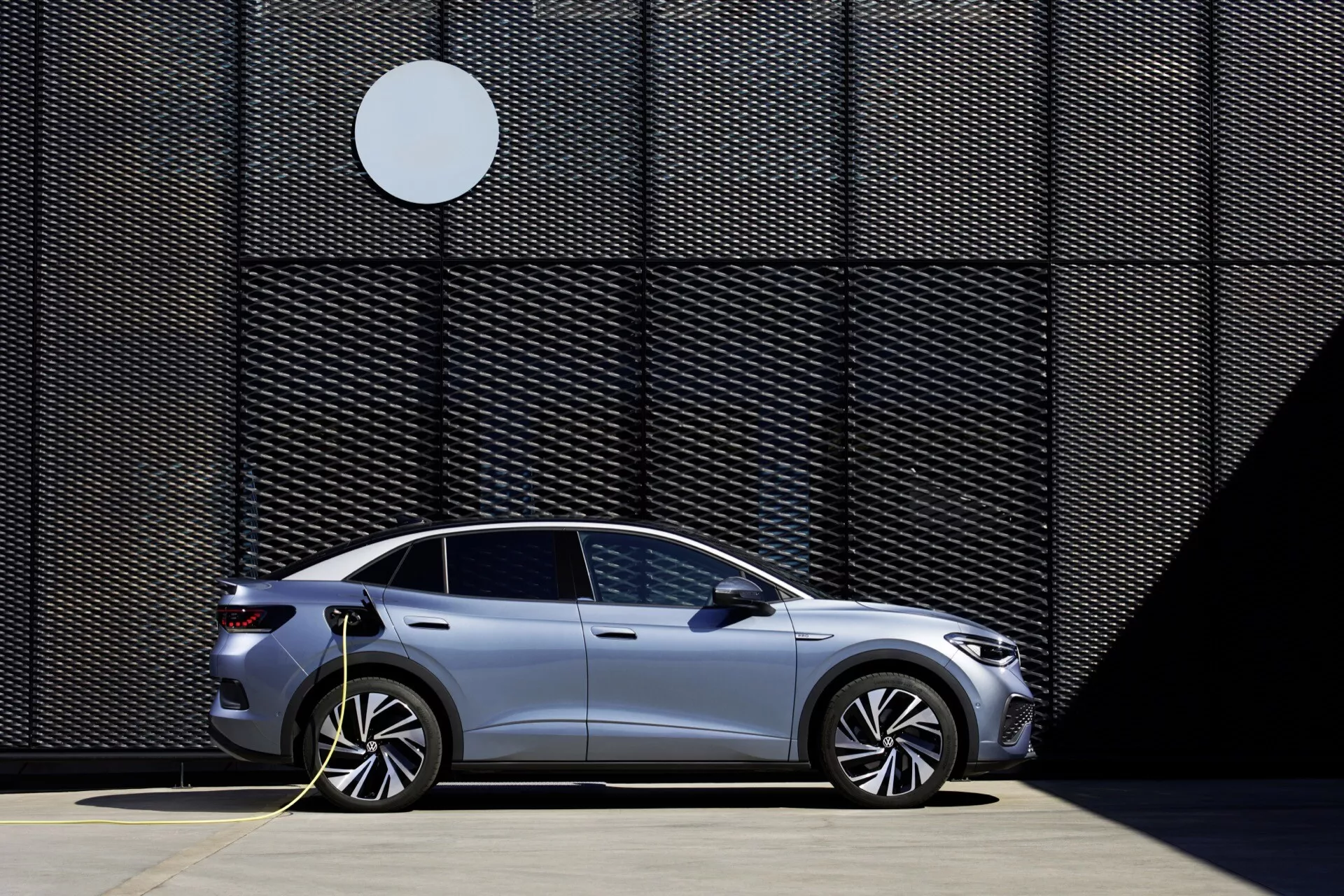 Boční profil stříbrného elektromobilu VW ID.5 2022 zaparkovaného a připojeného k nabíjecí stanici. Vozidlo má aerodynamická kola, čtyři dveře a moderní, elegantní design, vše zdůrazněno jasným, slunečným dnem. Na pozadí obrazu je geometrická stěna s kontrastním kruhovým tvarem, což zdůrazňuje umístění vozidla v určené oblasti pro nabíjení elektromobilů.
