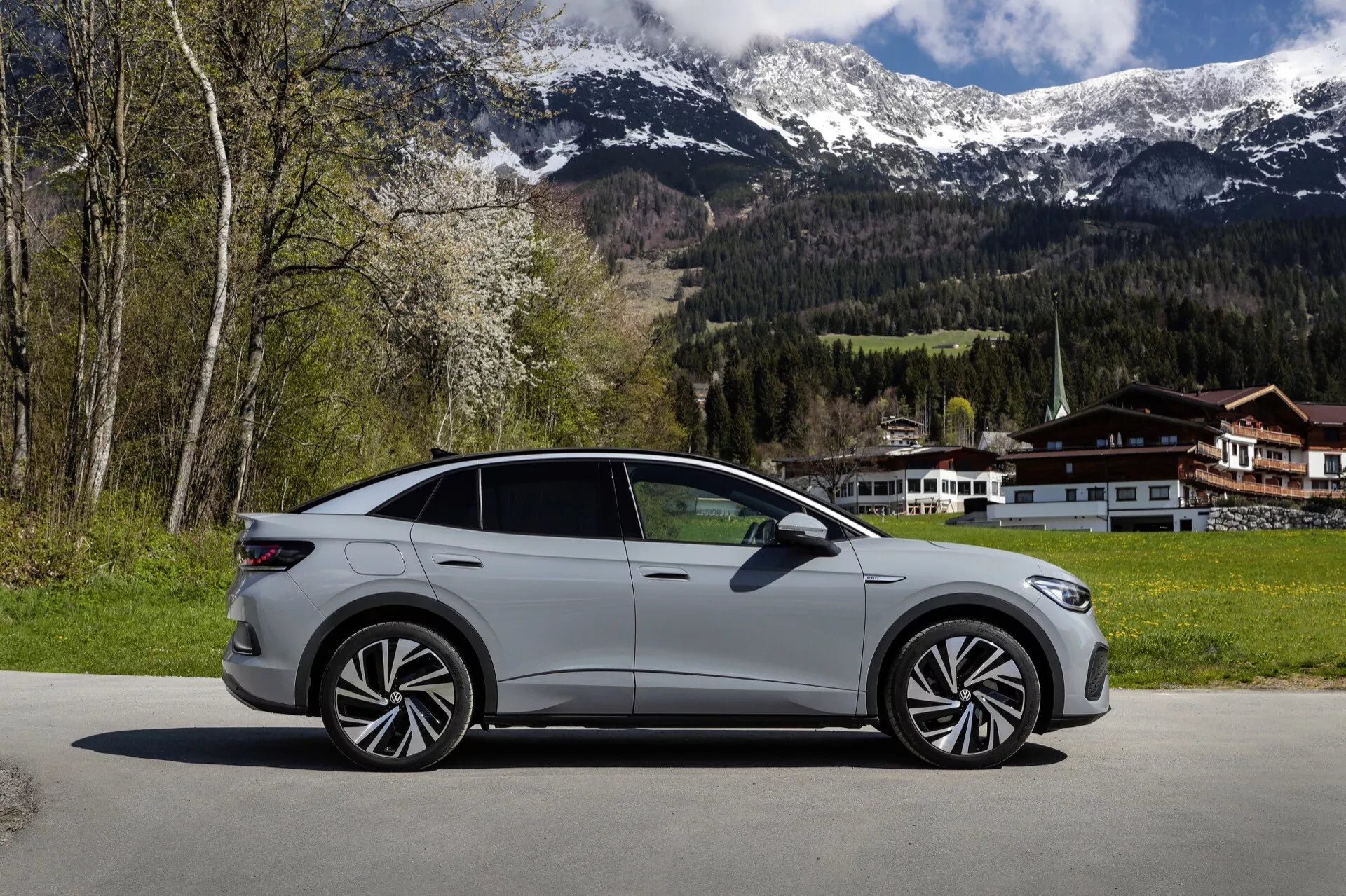 Boční profil stříbrno-šedého VW ID.5 z roku 2022 (1. generace), představující elegantní a aerodynamický design s výraznými obrysovými liniemi, moderními litými koly, barevnými bočními zrcátky a zarovnanými dveřními klikami, zaparkovaný na dlážděné ploše s malebným horským pozadím a domem ve stylu švýcarské chalupy.