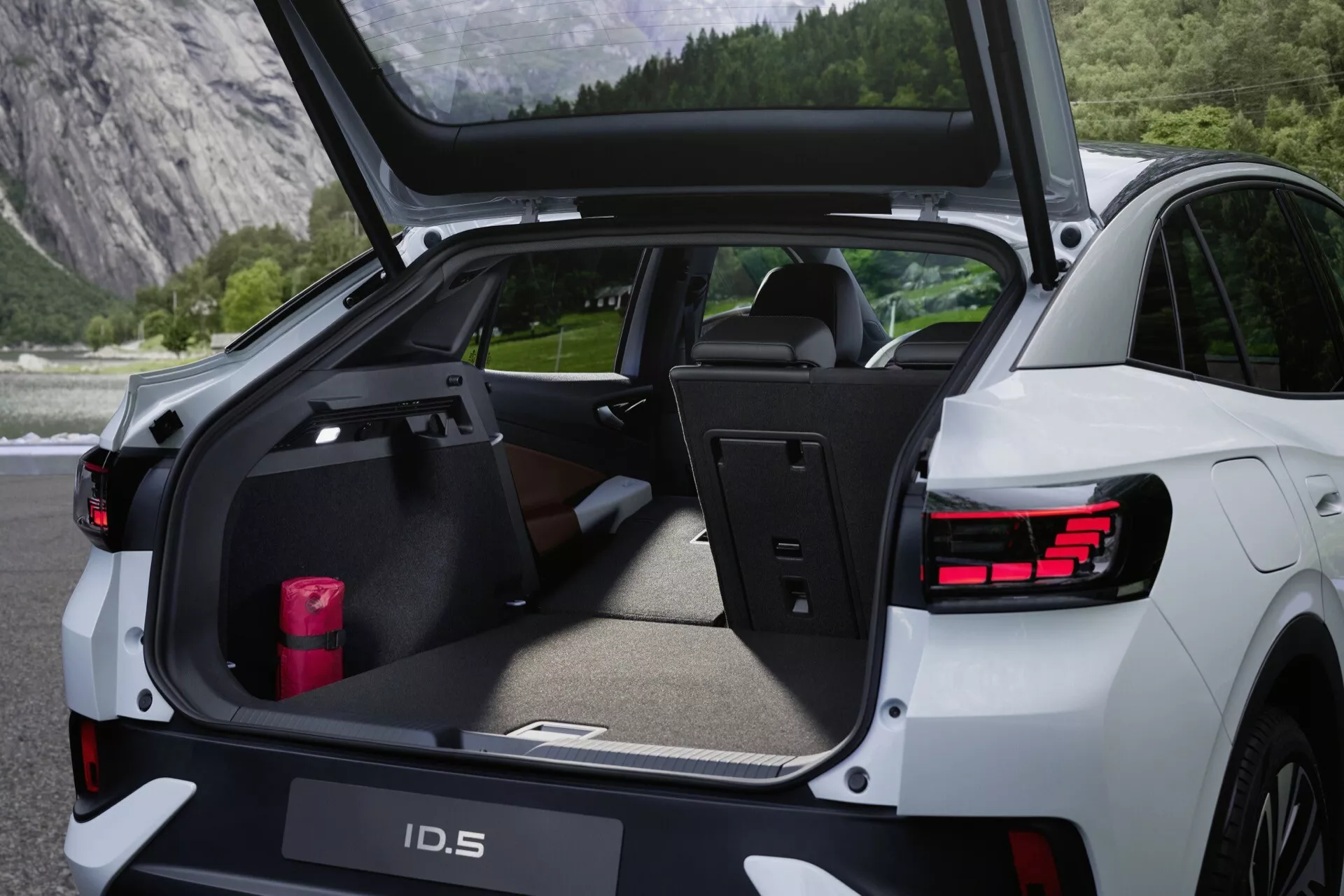 Zadní pohled na Volkswagen ID.5 2022 s otevřeným víkem kufru, ukazující zavazadlový prostor, zdůrazňující jeho praktičnost a kapacitu nákladu. Obrázek zvýrazňuje senzory na zadním nárazníku, LED zadní světla, elektrické víko kufru, vysoce kvalitní interiér, zadní sedadla s děleným sklopem, zatahovací kryt zavazadlového prostoru, plochý zavazadlový prostor s ochrannou podložkou a opěrku ruku s držáky na pohárky. 'ID.5' je viditelný na víku kufru.