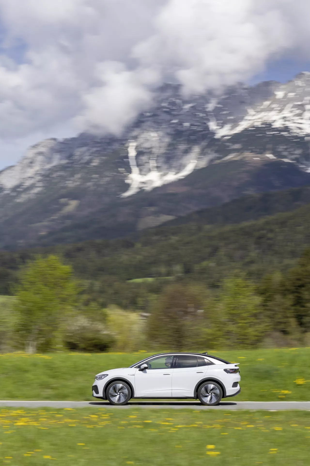 Dynamický boční profil VW ID.5 2022 v pohybu na malebné silnici s bujnou zelení a horskou kulisou, které ukazuje elegantní bílý design s litými koly, panoramatickou střechou a pokročilými funkcemi, který byl zachycen pomocí panning fotografie pro zdůraznění rychlosti.