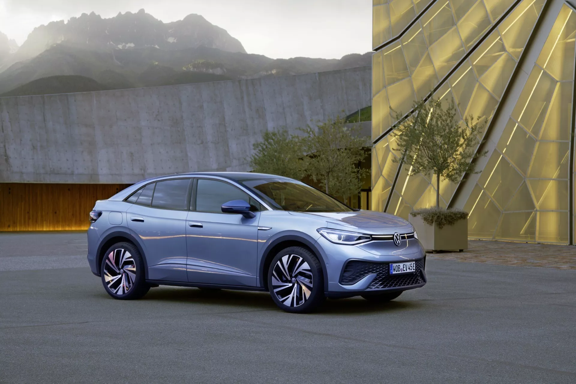 Tříčtvrteční přední pohled na první generaci 2022 VW ID.5 hatchbacku nebo crossover SUV. Obrázek zvýrazňuje levou stranu vozu zepředu, ukazující LED světlomety, minimalistickou přední masku, aerodynamický design kol a část kapoty, blatníku a čelního skla. Městské prostředí s geometrickými strukturami v pozadí dodává moderní kontext. Senzory na předním nárazníku naznačují pokročilé asistenční funkce řidiče.