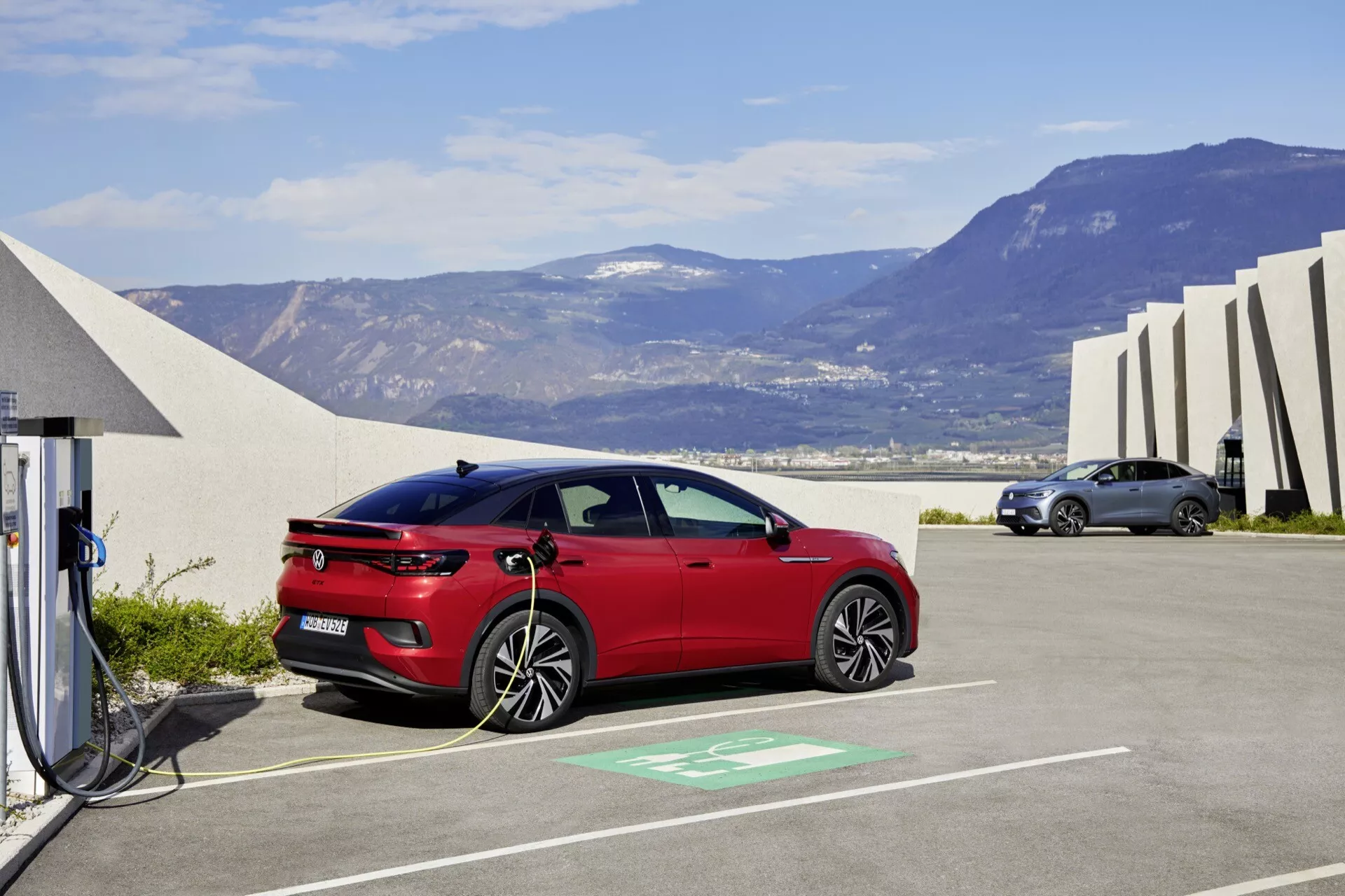 Obrázek ukazuje moderní červený hatchback VW ID.5 2022 zaparkovaný v nabíjecí stanici, což naznačuje, že se jedná o elektrický nebo plug-in hybridní vůz. Vozidlo je zobrazeno z profilu s jasným pohledem na jeho elegantní linie karoserie, aerodynamický design a otevřený nabíjecí port připojený k nabíjecí stanici viditelným kabelem. Kontext zahrnuje vyhrazené zelené parkovací místo, abstraktní architektonické struktury, otevřenou oblohu a horský terén, což zdůrazňuje ekologicky uvědomělé prostředí.