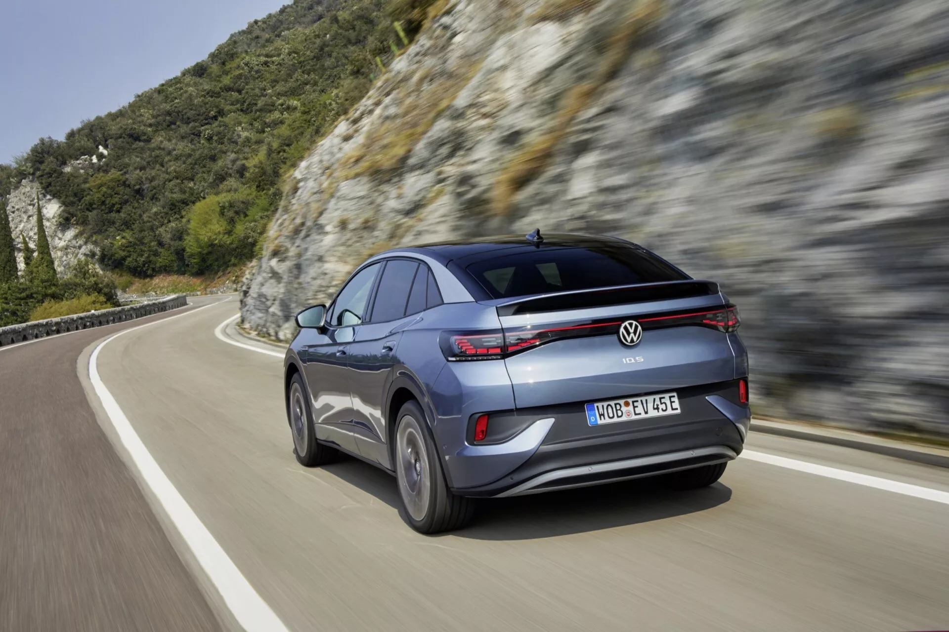 Moderní crossover SUV Volkswagen ID.5 2022 první generace v pohybu, zobrazený ze zadního tříčtvrtečního úhlu. Vozidlo je šedé nebo stříbrné s kovovým leskem, má ostrá, horizontální LED zadní světla, vícepaprsková litá kola a boční zpětné zrcátko stejné barvy jako karoserie s integrovaným směrovým světlem. Zadní nárazník obsahuje odrazky, senzory a zadní kameru. Snímek byl pořízen na horské nebo kopcovité silnici během dne a navozuje pocit rychlosti a pokročilého automobilového výkonu.