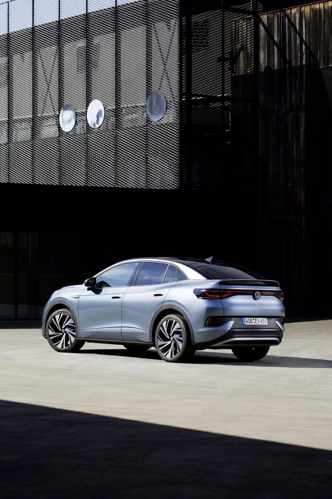 Obrázek sedanu VW ID.5 2022 (1. generace) z pohledu zezadu ze tří čtvrtin, ukazující jeho elegantní a moderní design s velkými hliníkovými koly, metalickou barvou, střechou sklopenou jako kupé, jemným spoilerem na víku kufru, tenkými LED zadními světly, zatmavenými okny a integrovanými výfukovými otvory, vše zasazeno do městského prostředí s geometrickými architektonickými prvky na pozadí.