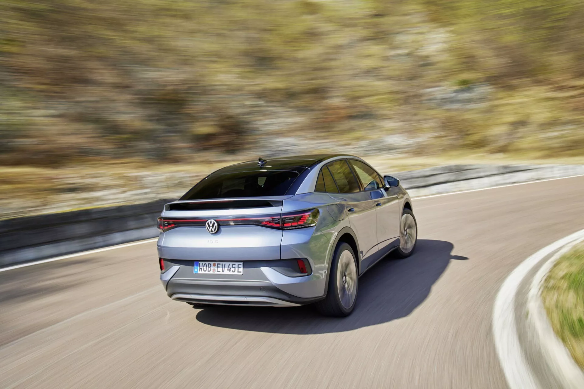 Zadní tříčtvrteční pohled na jedoucí VW ID.5 2022 (1. generace) za denního světla, zachycený z nižšího úhlu u zadního kola. Obrázek zvýrazňuje křivkovou svahovitou linii střechy, horizontální zadní světelné svazky, centrálně umístěné logo výrobce pod integrovaným zadním spoilerem, centrálně umístěnou registrační značku, anténu ve tvaru žraločí ploutve a střešní okno/měsíční okno. Pohyb vozidla je naznačen rozmazaným pozadím a zkreslením kol, což naznačuje střední až vysokou rychlost, a odrazivými povrchy laku a zadního skla, což naznačuje slunečný den.