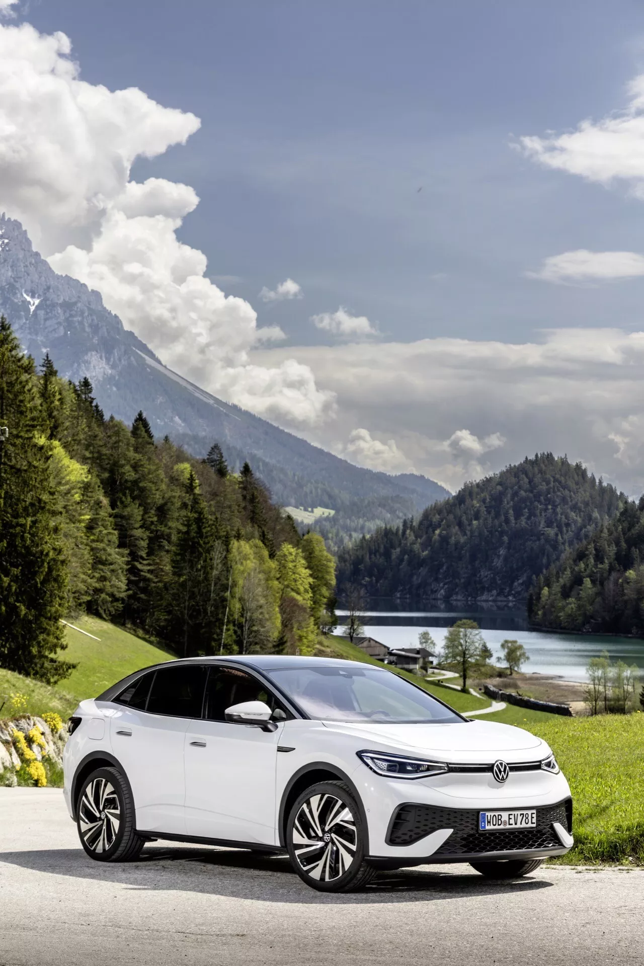 Obrázek moderního elektrického hatchbacku, VW ID.5 2022 (1. generace), umístěného na silnici s scenickým pozadím jezera a hor. Auto je zachyceno z úhlu tři čtvrtiny zepředu, ukazující jeho aerodynamická litá kola, LED denní světla, design světlometů a hladké linie karoserie. Přirozené osvětlení a malebné prostředí zdůrazňují ekologický aspekt vozidla, s dostatečným prostorem kolem něj pro pocit otevřenosti.