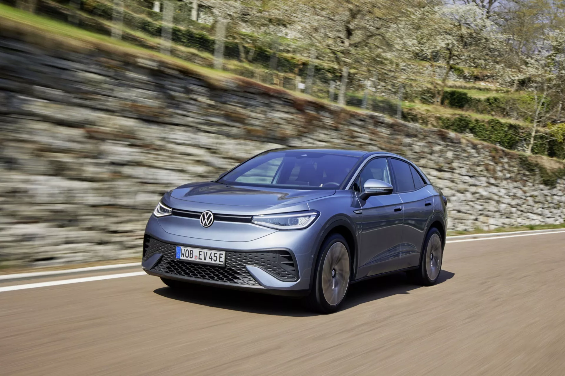 Dynamický pohled na Volkswagen ID.5 z roku 2022 (první generace) zachycený z nízkého, předního tříčtvrtečního úhlu, zobrazující čelní nárazník, mřížku, kapotu, čelní sklo a profil na straně řidiče. Auto, natřené v metalické barvě, se pohybuje po dlážděné silnici, čímž předvádí svůj elegantní, aerodynamický design a technologii elektrického vozidla. Čelní část, slitinová kola a značka výrobce jsou jasně viditelné, zdůrazňující moderní osvětlovací technologií a uzavřenou masku chladiče typickou pro elektrická vozidla.