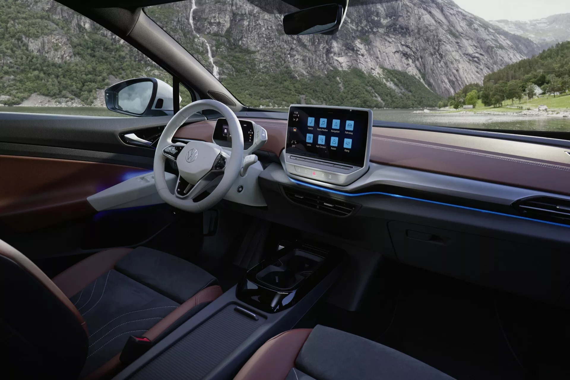 Interiér VW ID.5 2022. Kamerový úhel zajišťuje pohled ze zadního pravého sedadla spolujezdce, ukazující elegantní a minimalistický design a luxusní dojem. Vlevo umístěný volant postrádá tradiční přístrojový panel, což naznačuje digitální displej. Ve středu palubní desky výrazný dotykový displej zdůrazňuje pokročilé infotainmentové funkce. Minimální fyzické ovládací prvky a automatická převodovka svědčí o moderních designových preferencích. Palubní deska a dveřní panely zdobí dřevěné nebo dřevěně vypadající materiály a tmavé plasty, posilující prémiový vzhled. Přírodní světlo z velkého bočního okna rozjasňuje interiér, který má tmavé čalounění s červenými akcenty.