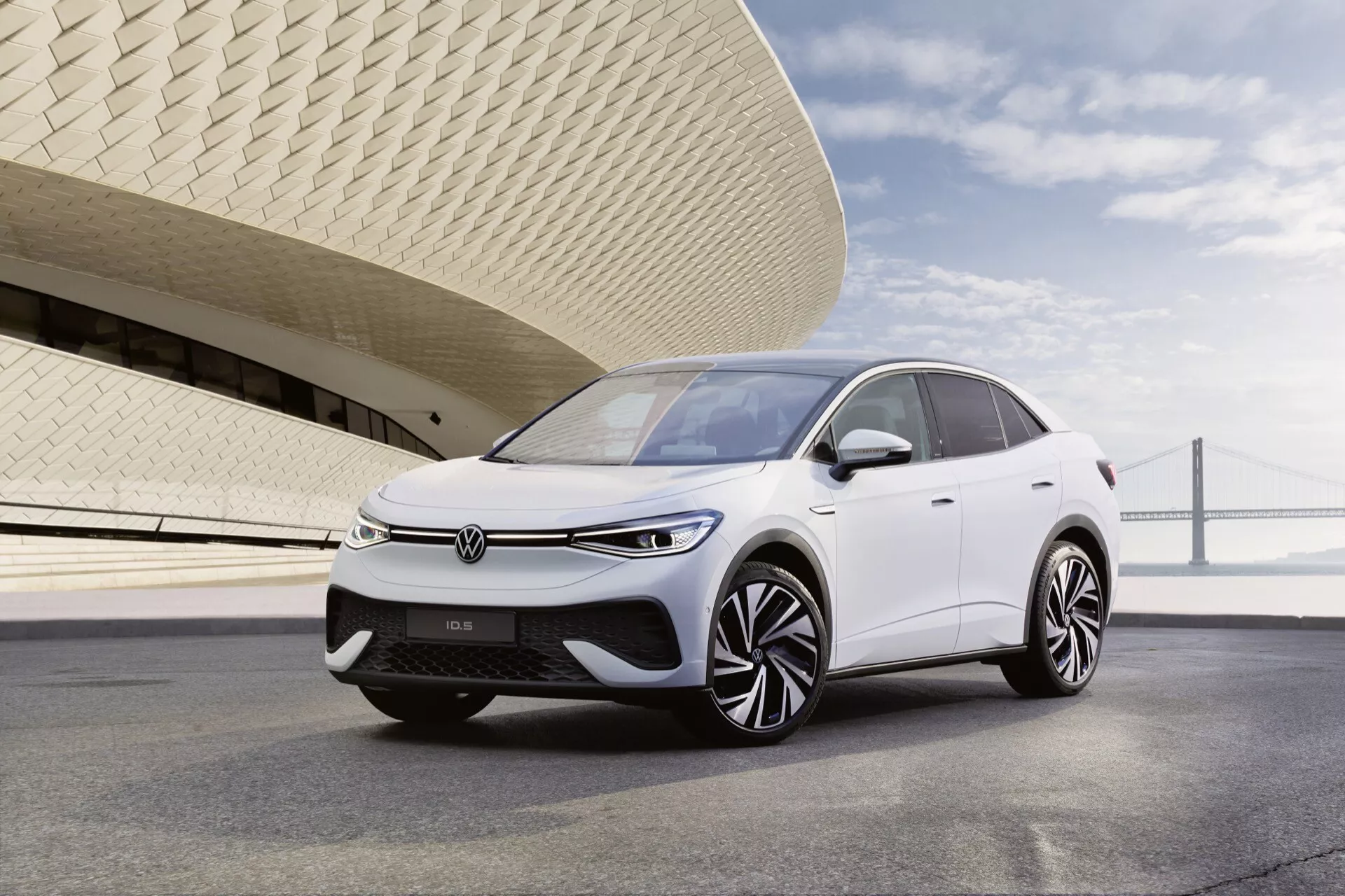 Obrázek Volkswagen ID.5 z roku 2022 (první generace) v bílé barvě, umístěný v tříčtvrtečním úhlu, aby vynikl přední a řidičův bok. Pohled zahrnuje přední mřížku, elegantní LED světlomety, aerodynamickou kapotu a propracovaná litá kola. Zvýšený postoj auta a uzavřená mřížka naznačují, že jde o elektrické vozidlo (EV) s pokročilými funkcemi, pravděpodobně včetně senzorů pro asistenční technologie řízení. Auto je zobrazeno za denního světla, zaparkované na hladkém dláždění s moderní budovou v pozadí, což zdůrazňuje jeho současný a efektivní design.