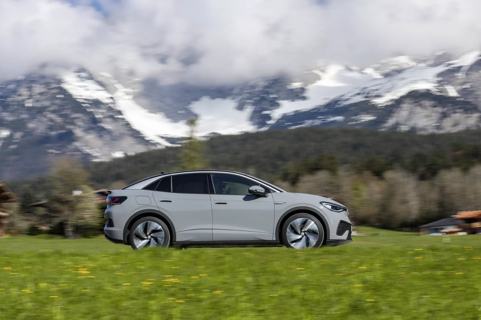 Dynamický snímek moderního VW ID5 2022 (první generace) v pohybu, zachycený technikou panorámování, která zobrazuje rychlost a pohyb. Pohled z bočního profilu s aerodynamickými liniemi, nízkoprofilovými pneumatikami a moderním designem kol. Vůz, pravděpodobně sportovní sedan s kupé siluetou a velkými okny možná včetně panoramatické střechy, je zaostřen, když se pohybuje zleva doprava proti rozmazanému pozadí. S propláklými klikami dveří a bočními zrcátky, nastavení naznačuje venkovské či malebné prostředí s travnatým terénem a horami v pozadí.