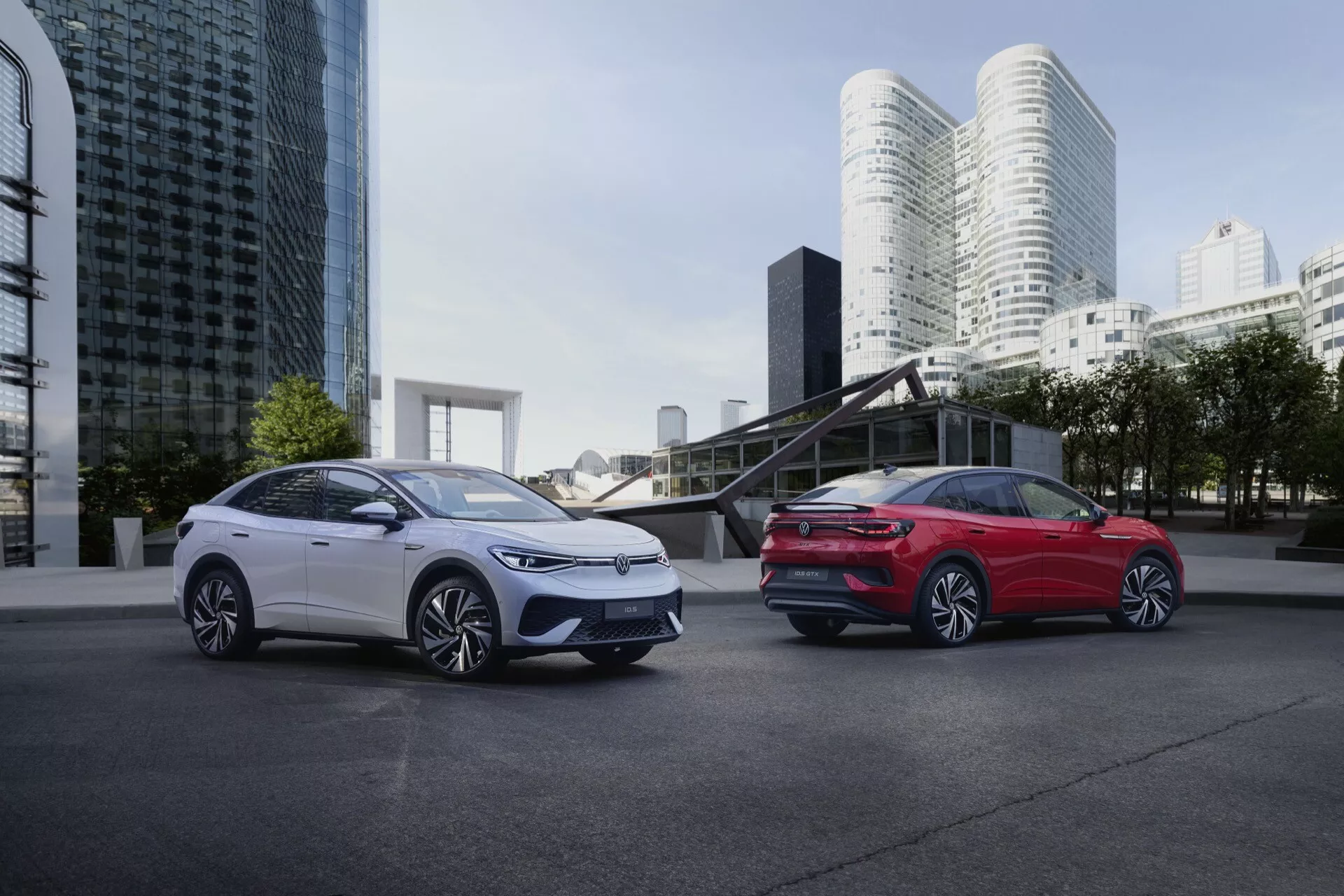 Dva elegantní a moderní elektrické nebo hybridní vozy VW ID.5 2022 (1. generace) postavené na pozadí městské scenérie s moderní architekturou. Obrázek, pořízený z nízkého úhlu, zdůrazňuje rozměry vozidel. Bílý vůz vlevo ukazuje svou přední levou stranu, včetně mřížky, designu světlometů, kontur kapoty a kola na straně řidiče. Červený vůz vpravo ukazuje svou zadní pravou stranu s viditelným designem zadních světel, zadním nárazníkem a křivkou zadního čtvrtpanelu. Oba vozy mají aerodynamická kola z lehkých slitin s komplikovanými vzory paprsků a jejich lesklý povrch odráží okolní světlo raného rána nebo pozdního odpoledne.