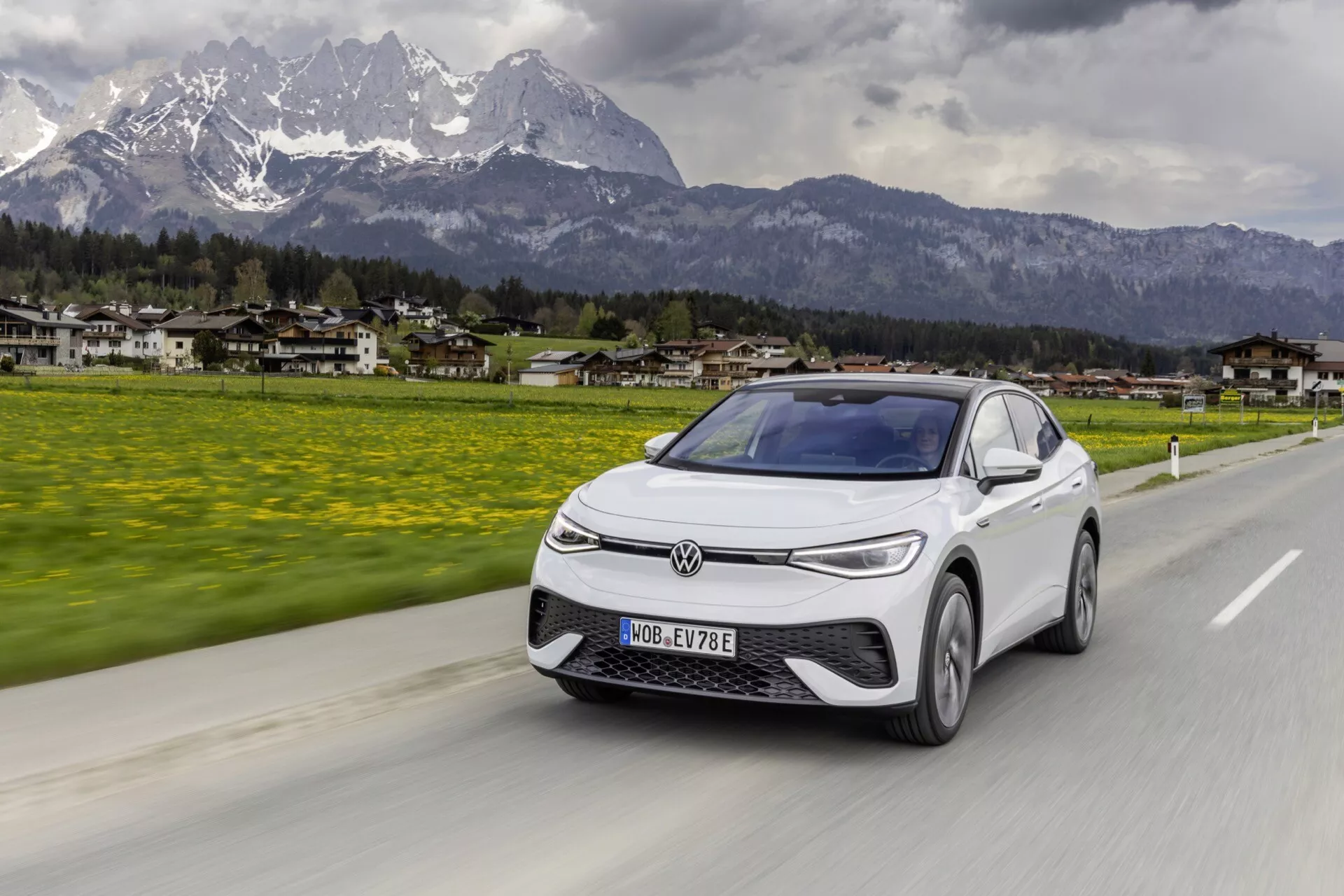 Obrázek zobrazuje vůz VW ID.5 2022 první generace jedoucí po silnici s malebným pozadím hor a malého města. Záběr z tříčtvrtinového předního úhlu se zaměřuje na přední levou část, ukazuje mřížku, světlomety, kapotu, čelní sklo a část levých dveří a okna řidiče. Elegantní, aerodynamický elektromobil má zavřenou mřížku, úzké hranaté LED světlomety, pokročilé aerodynamické linie a moderní ráfky na kolech v pohybu. Vůz je zobrazen v pohybu, s integrovanými ukazateli na bočních zrcátkách a panoramatickým čelním sklem táhnoucím se směrem ke střeše, což naznačuje zlepšenou viditelnost řidiče.
