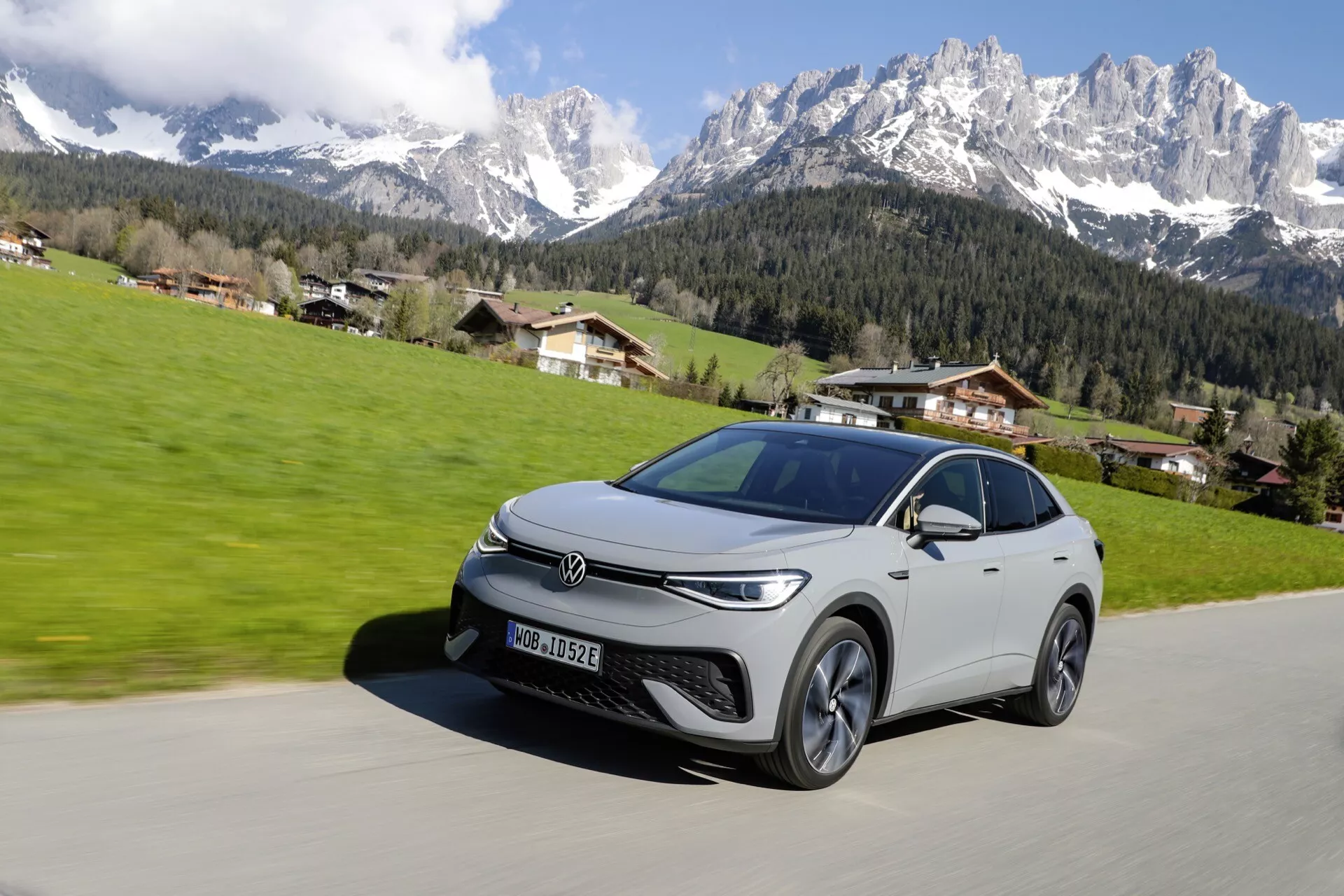 Obrázek elegantního stříbrného crossoveru SUV Volkswagen zachyceného za jasného slunečného dne s horskou krajinou a jasnou oblohou na pozadí. Auto je viděno z nízkého úhlu, který ukazuje přední levý bok, s ostrými světlomety, úzkou maskou chladiče, vícepaprskovými slitinovými koly a elegantními liniemi naznačujícími moderní, možná elektrický nebo hybridní design. Auto je na dlážděné silnici a vypadá to, že se pohybuje proti klidnému venkovskému pozadí.