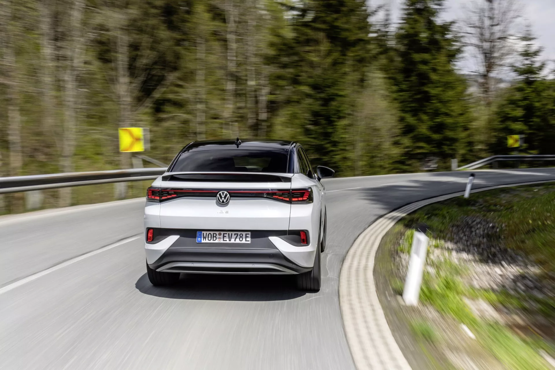 Dynamický pohled na záď jedoucího vozu VW ID5 2022 (1. generace) na zakřivené silnici, viděný z nízko-střední výšky se zaměřením na moderní designové prvky, jako je výrazný horizontální světelný pás, rozpoznatelný emblém a označení modelu. Registrační značka naznačuje, že se jedná o elektrické vozidlo s označením 'E'. Viditelné parkovací senzory a absence výfukových trubek ukazují na pokročilé funkce, jako je parkovací asistenční technologie a elektrický pohon.