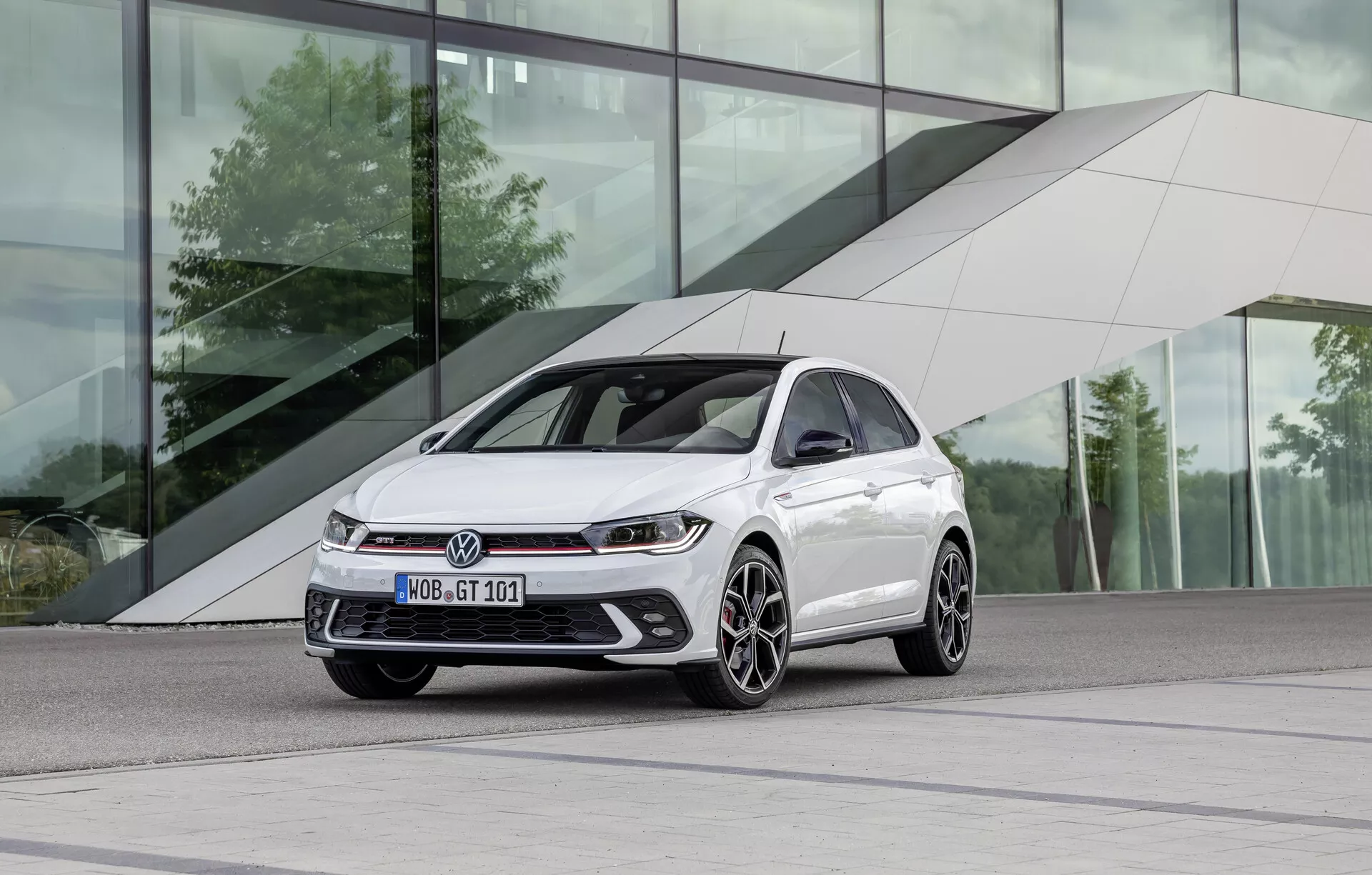 Moderní hatchback auto v lesklé bílé barvě umístěné na levé straně rámu, focené z nízkého, tříčtvrtečního předního úhlu. Elegantní a sportovní vnější design zahrnuje skloněnou kapotu, dynamické linie, ostré LED světlomety, výraznou přední mřížku, integrované mlhové světlomety, výkonnostní nasávací otvory a vícepaprsková litá kola s dvou tónovým provedením. Auto je zasazeno do městského prostředí s moderní budovou s elegantními designovými rysy a reflexním sklem.