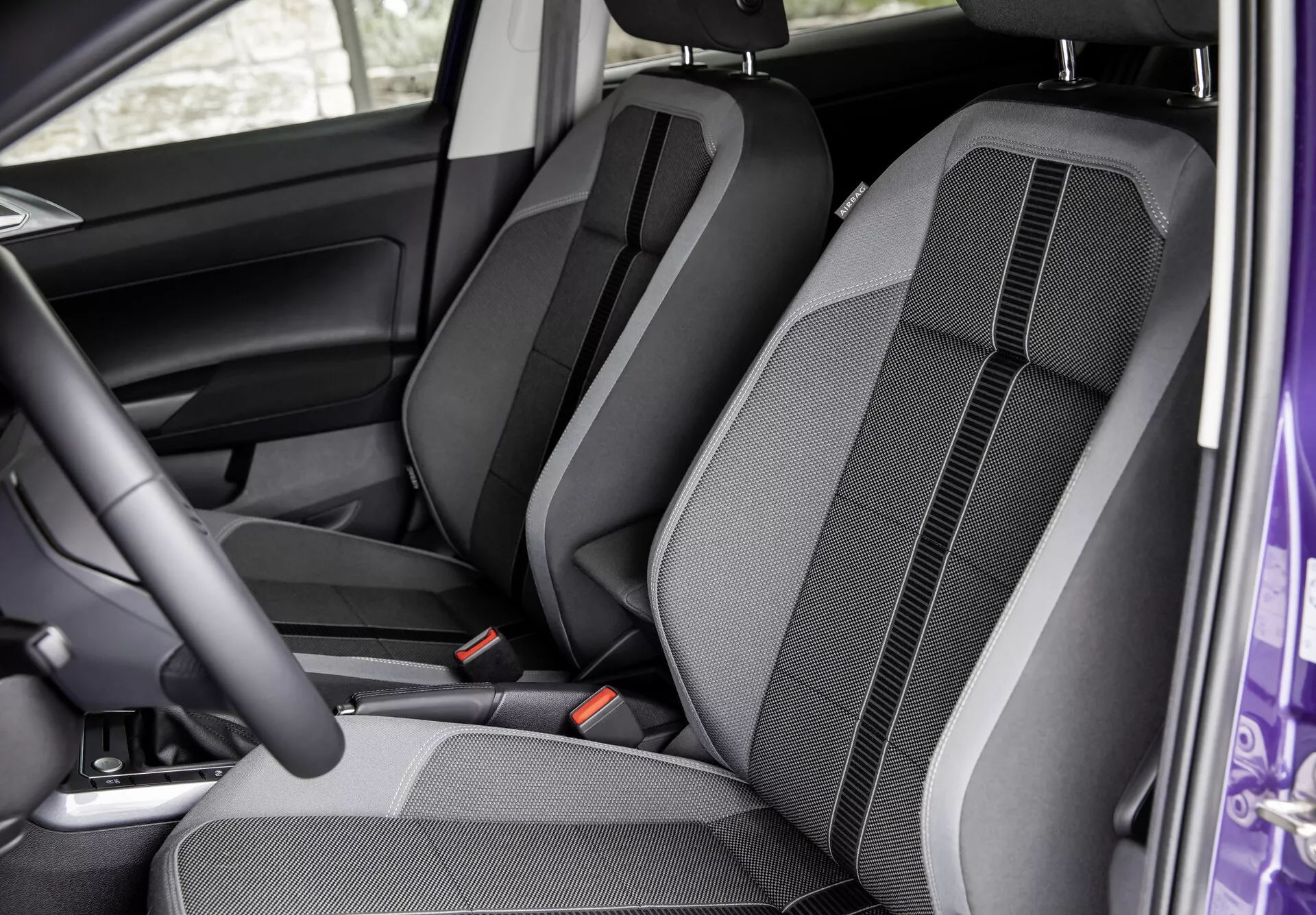 Interiér VW Polo 2021 (VI-facelift) z pohledu otevřených předních dveří na sedadla předních a zadních sedadel. Zobrazena je konfigurace sedadel, design látky, prošívání a detaily tvarování. Viditelné jsou výškově nastavitelné opěrky hlavy, bezpečnostní pásy a částečný pohled na dveřní panel u řidiče, zdůrazňující komfort a interiérový design.
