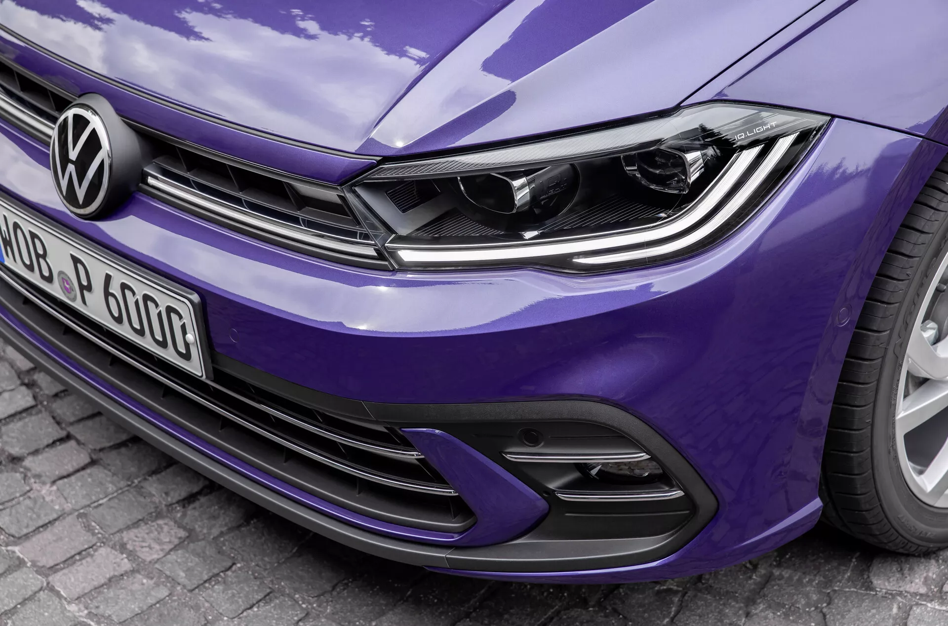 Detailní, šikmý přední pohled na modrý Volkswagen Polo 2021 VI-facelift, který ukazuje moderní design světlometů s integrovanými LED denními světly, výrazným logem Volkswagen na horizontálních mřížkách a sportovním nárazníkem s přívody vzduchu a designem z medové mřížky. Detaily zdůrazňují elegantní linie kapoty a lesklý povrch laku, což naznačuje pokročilou osvětlovací a potenciální asistenci řidiče.