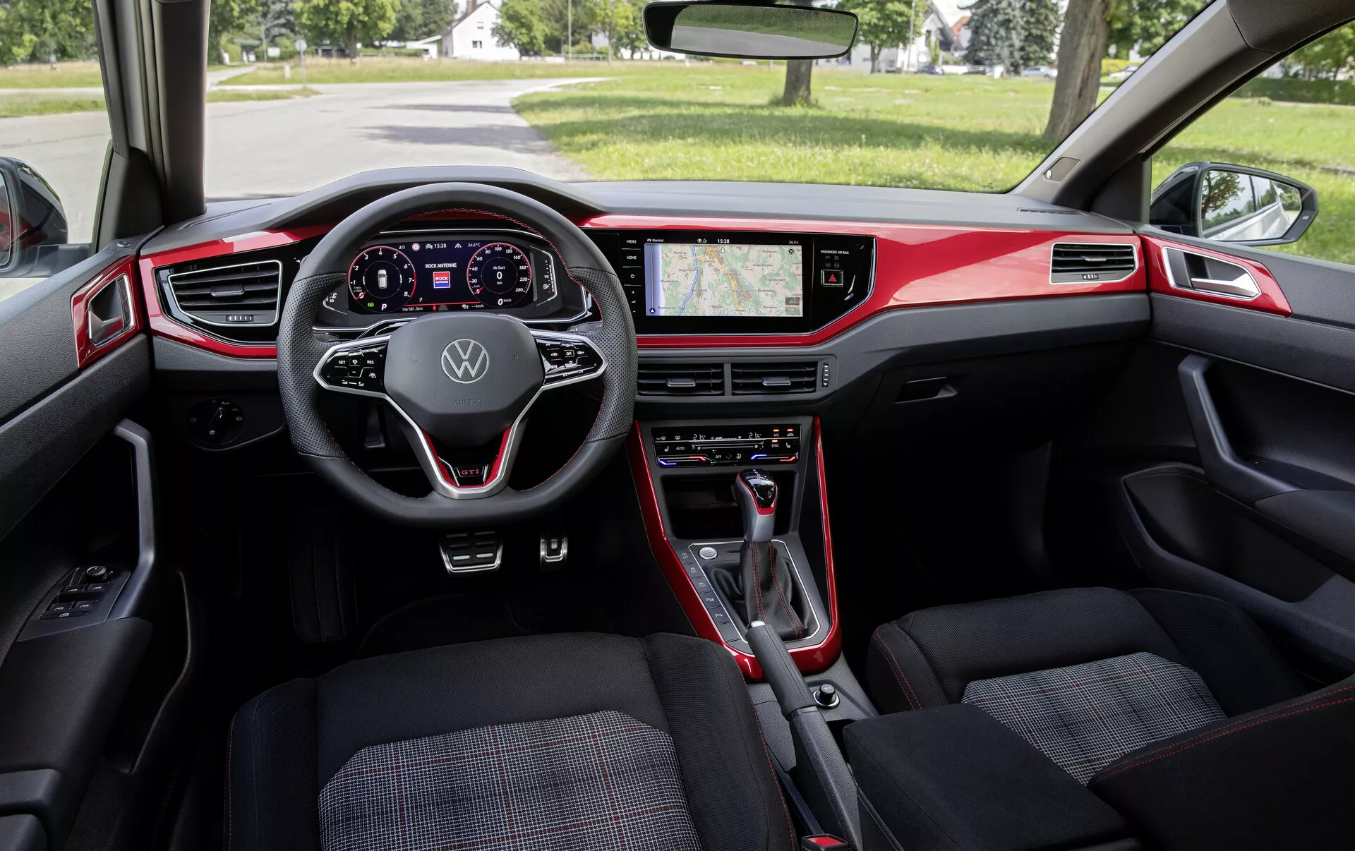 Pohled do interiéru VW Polo 2021 (VI-facelift) z perspektivy zadního sedadla. Širokoúhlý záběr zachycuje palubní desku, přední sedadla a zpětné zrcátko. Mezi výrazné prvky patří moderní multifunkční volant s logem Volkswagen, plně digitální přístrojový štít, velký dotykový infotainment displej zobrazující GPS navigaci a fyzické ovládání klimatizace. Převážně černý interiér s červenými akcenty zdůrazňuje sportovní design. Přední sedadla mají vzorované čalounění a volič převodovky naznačuje automatickou převodovku. Celkově interiér spojuje funkčnost s moderním designem a pokročilou technologií.