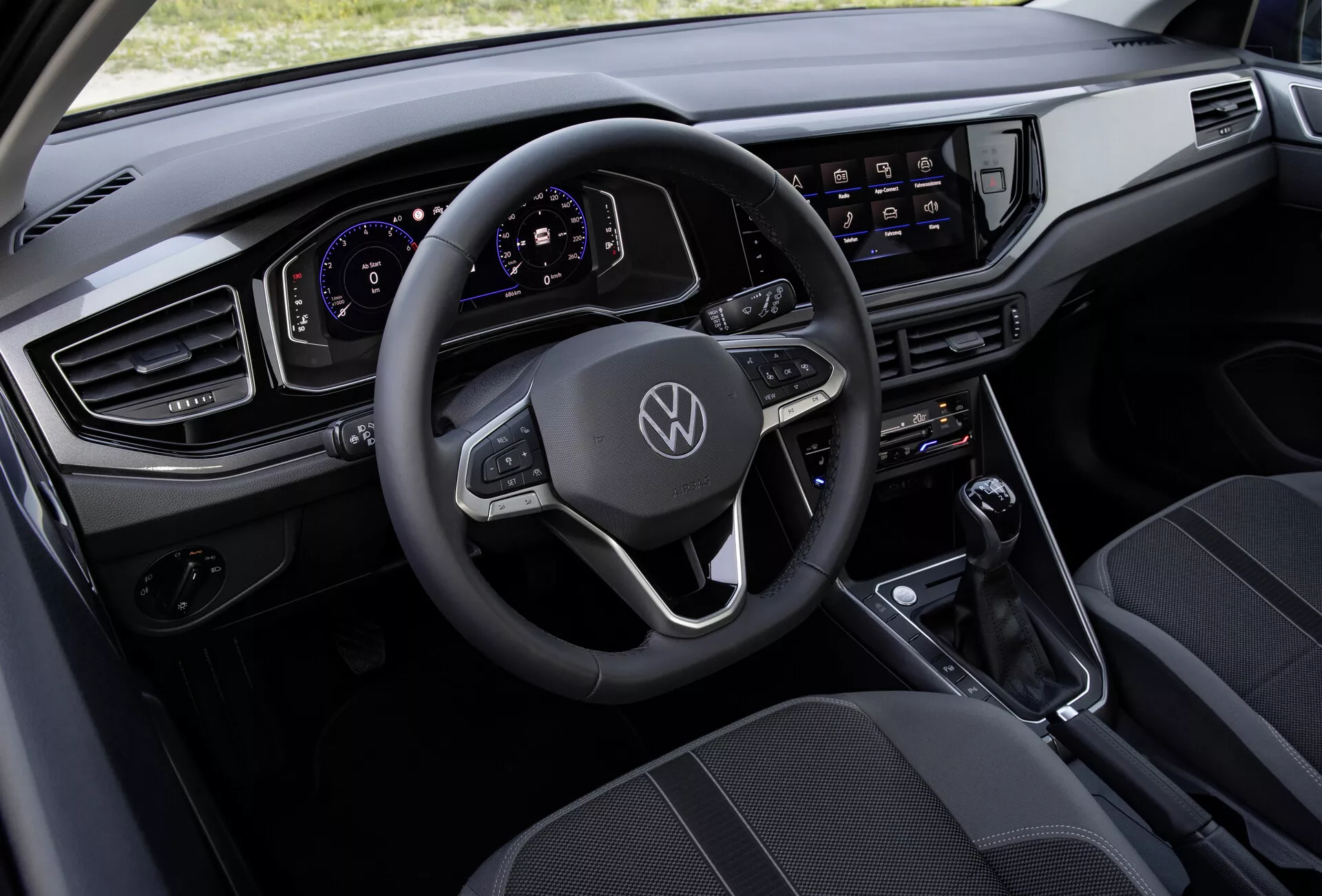Pohled z výšky na interiér VW Polo 2021 (VI-facelift), který ukazuje multifunkční kožený volant s plochým spodkem zdobený logem Volkswagen, plně digitální a přizpůsobitelný přístrojový štít, velký dotykový infotainment systém, moderní vodorovnou palubní desku s elegantními, hranatými větracími otvory, minimalistickou středovou konzoli s řadicí pákou pro automatickou převodovku a kvalitní látková sedadla s vzorovaným designem.