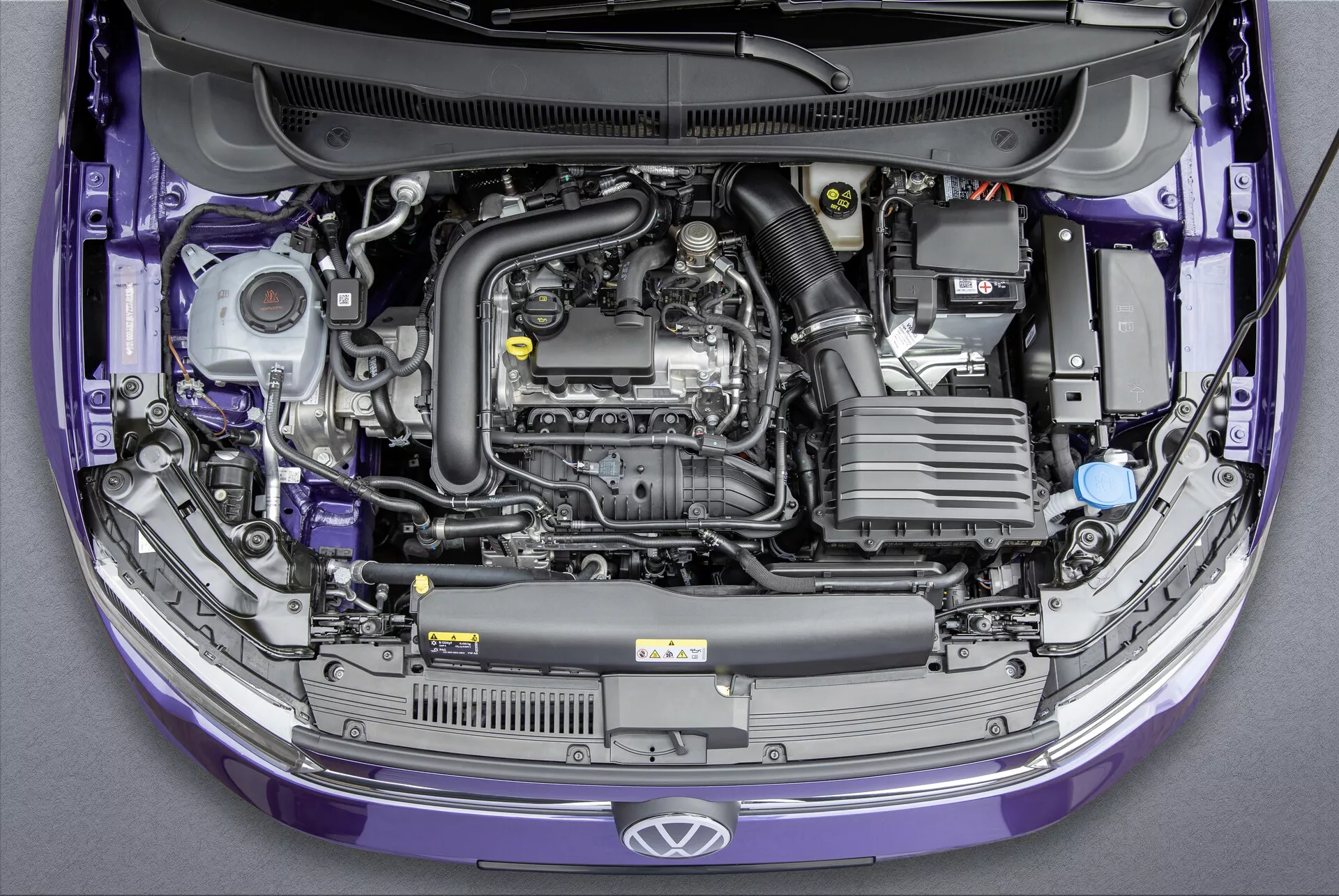 Pohled shora na Volkswagen Polo 2021 (VI-Facelift) se zvednutou kapotou, který odhaluje příčně umístěný motor a různé komponenty v motorovém prostoru. Obrázek zvýrazňuje plastový kryt motoru, baterii, systém sání vzduchu, nádržku chladicí kapaliny s modrým víčkem, různé nádoby na kapaliny, součásti motoru s kovovým a gumovým potrubím a pojistkovou skříňku. Karoserie auta je fialová a ve standardním továrním stavu. Ideální pro kategorie týkající se automobilových motorů, údržby vozidel, automobilového inženýrství a výroby automobilů.