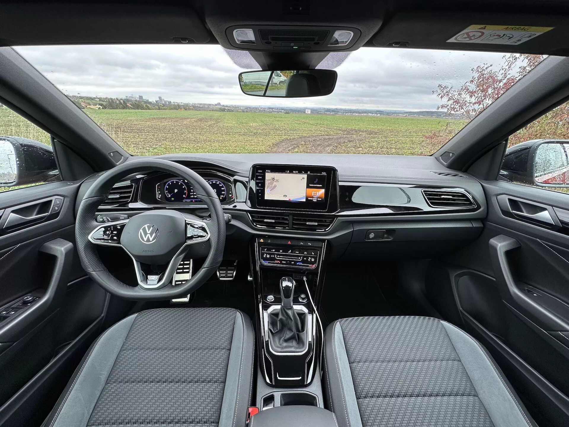 Interiérový pohled zezadu dopředu na VW T-Roc Cabriolet 2022 ukazující aerodynamický palubní desku s chromovými akcenty, digitální obrazovku infotainmentu, pokročilé klimatizace s duálním ovládáním, kožený volant s multifunkčními ovládáními a logem Volkswagen, řadicí páku automatu, a moderní sedadla s dvoubarevnou textilní tapicerií a prošívaným vzorem.
