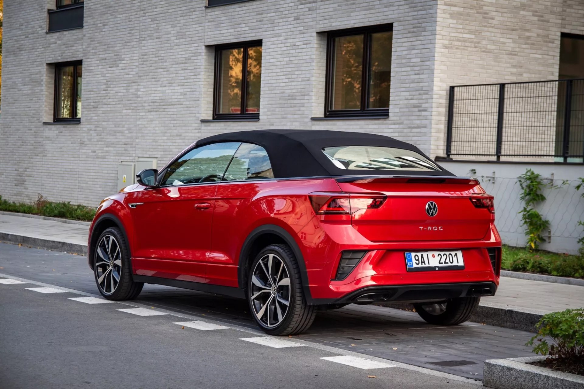 Zadní tříčtvrteční pohled na červený Volkswagen T-Roc Cabriolet zaparkovaný na ulici, zdůrazňující jeho černou skládací střechu, elegantní LED zadní světla, zadní spoiler, dvojité výfukové koncovky a integrované parkovací senzory na pozadí světle zbarvené cihlové budovy.