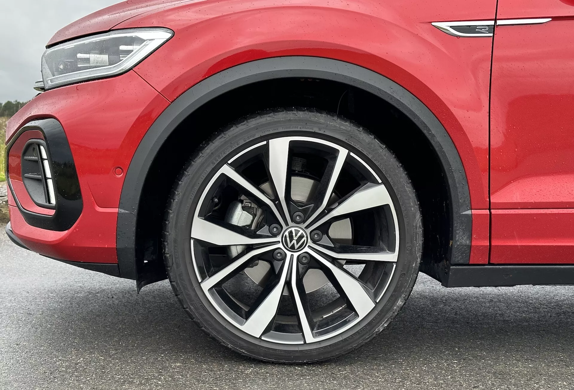 Detailní pohled na VW T-Roc 2022 se zaměřením na slitinové kolo s vícepaprskovým designem a dvoubarevným provedením, nízkoprofilovou pneumatiku a část předního blatníku ukazující sportovně červené karoserie a detail LED osvětlení