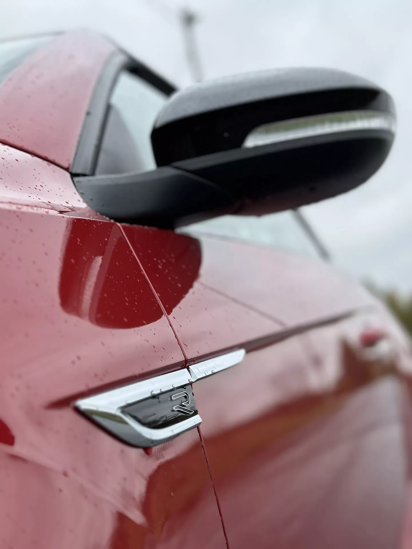 Detailní boční pohled na červený VW T-Roc Cabriolet 2022, zdůrazňující zadní část s detailním pohledem na černé boční zrcátko, kliku dveří s bezklíčovým vstupem a designové prvky auta včetně odznaku, pod mírným vzhůru směřujícím úhlem s kapkami vody na povrchu naznačující nedávný déšť nebo mytí, proti rozmazanému pozadí