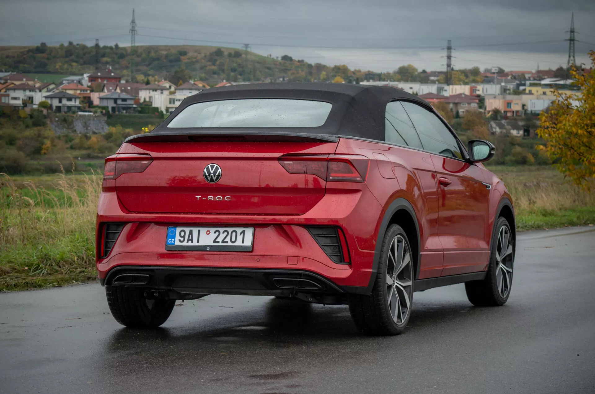 Červený Volkswagen T-Roc Cabriolet 2022 ve tříčtvrtečním zadním pohledu na mokré silnici, prezentující zadní design, boční profil, design kol, LED zadní světla, zadní nárazník s integrovanými senzory, dvojité výfukové trubky, slitinová kola s více paprsky a skládací střechu