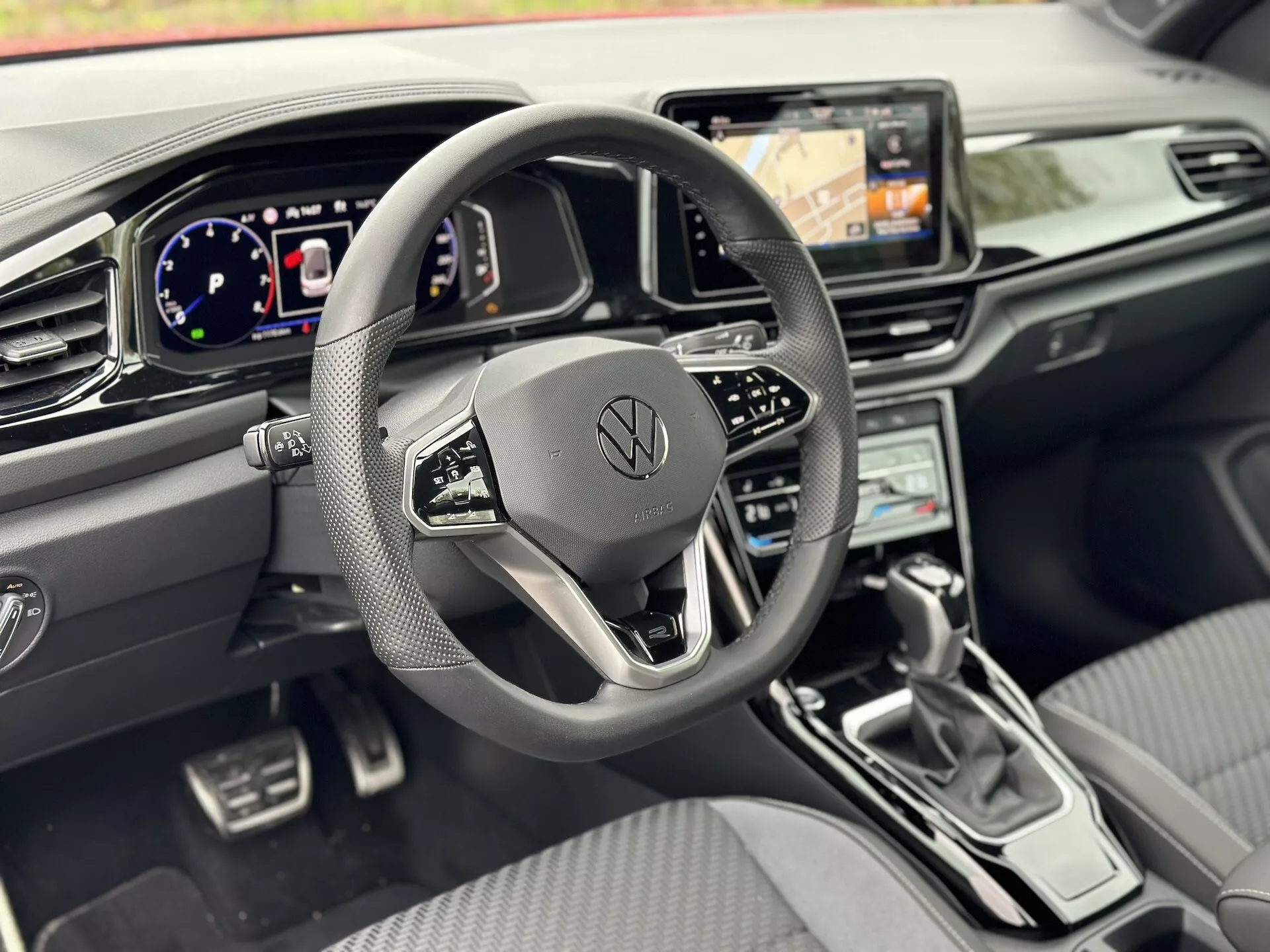 Interiér Volkswagen T-Roc Cabriolet 2022 z pohledu řidiče s důrazem na tříramenný volant s multifunkčními tlačítky a logem Volkswagen, digitální palubní desku, infotainment systém s navigací, klimatizaci, elegantní řadicí páku, ovládání asistenčních systémů a prémiový design kokpitu s kovovými akcenty a koženkovými sedadly.