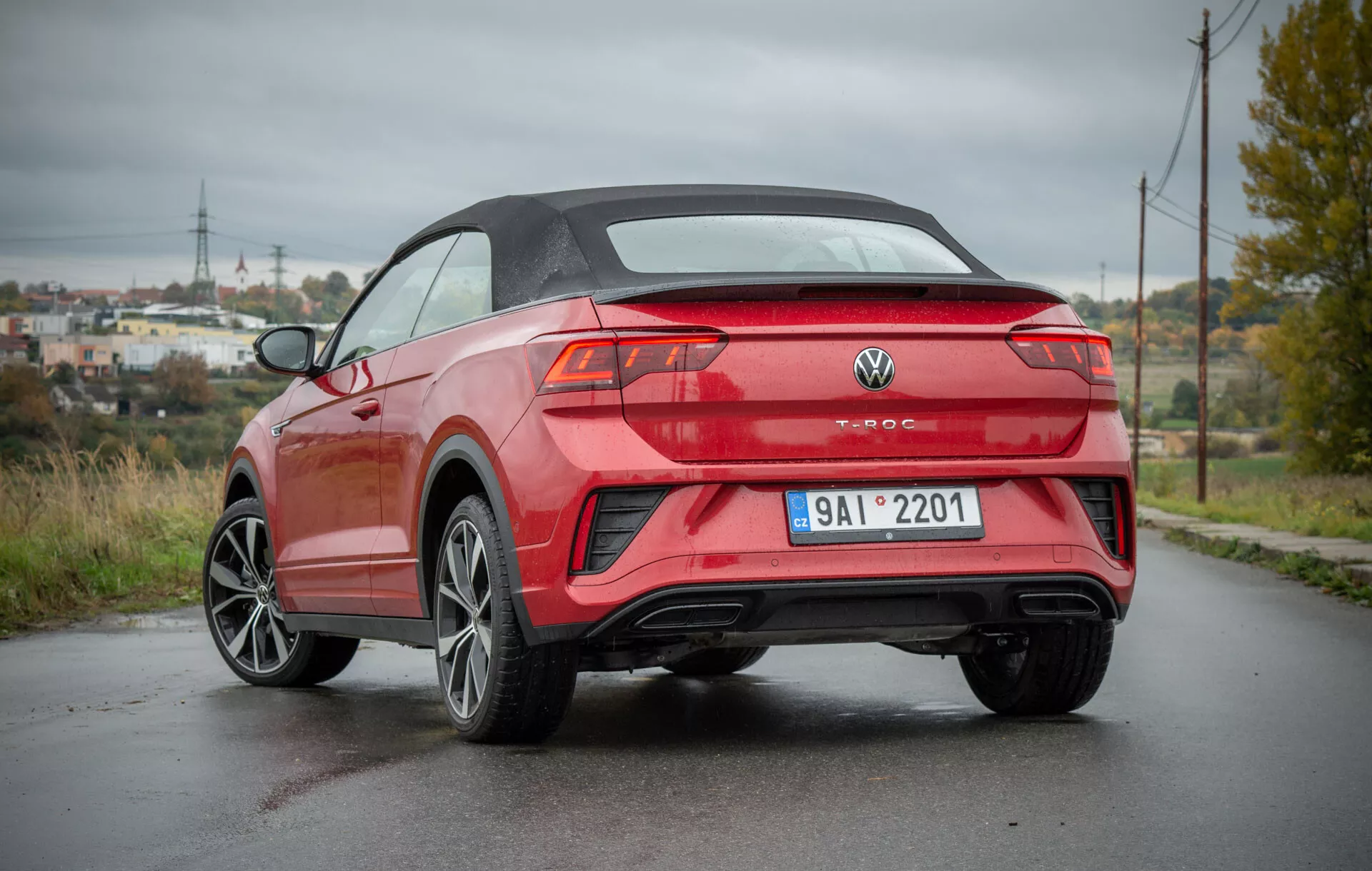 Zadní trojčtvrtě pohled na VW T-Roc Cabriolet 2022 zaparkovaný na vlhké silnici, který předvádí jeho moderní kabrioletní design, ostré linie karoserie, sportovní slitinová kola, elegantní zadní světla a dvojité výfuky, na pozadí částečně venkovské krajiny s elektrickými pylony a bujnou zelení.