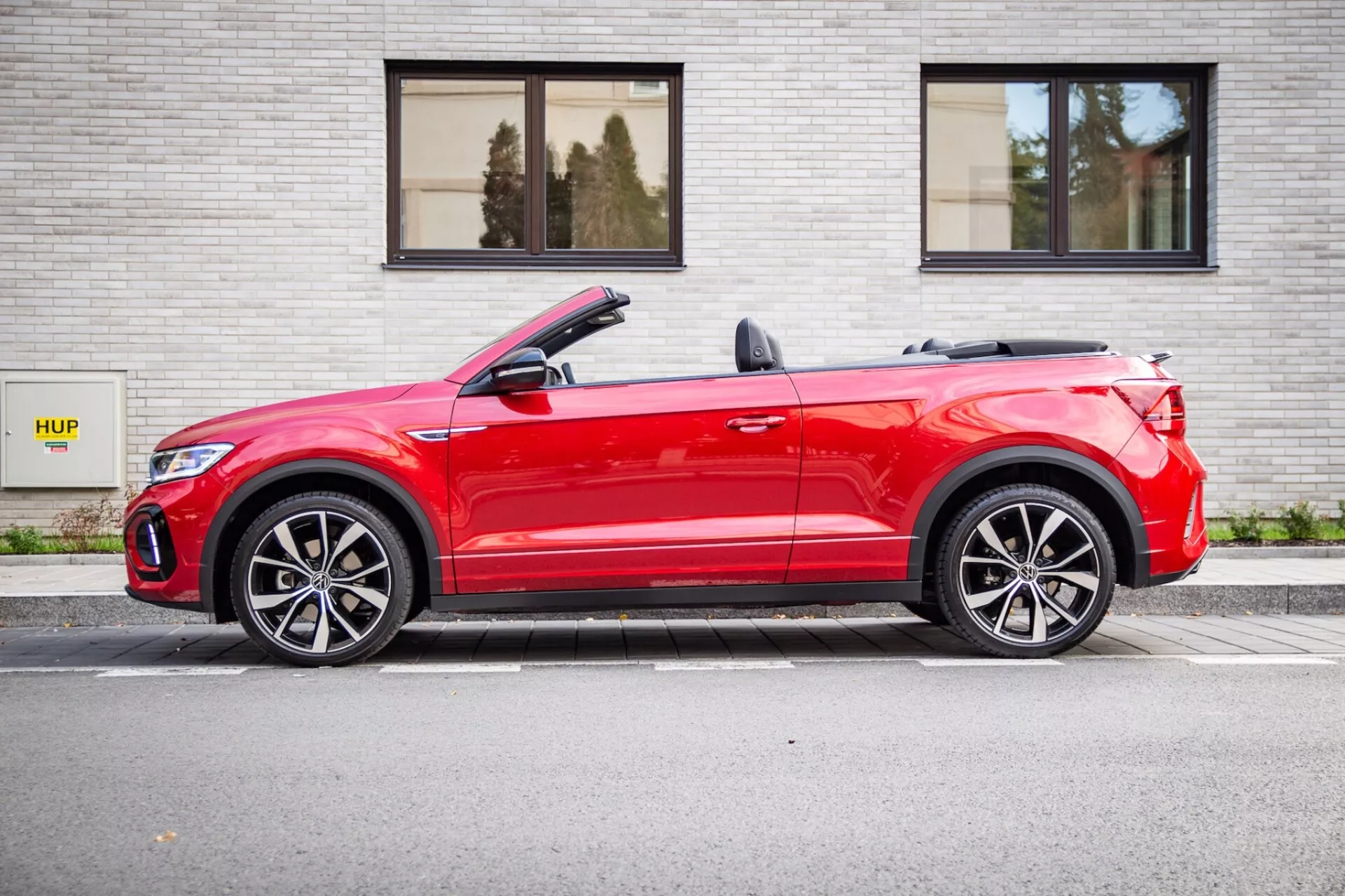 Červený kabriolet Volkswagen T-Roc 2022 zaparkovaný rovnoběžně s šedou cihlovou zdí s nápisem HUP, zobrazuje boční profil s mírným úhlem zepředu, stahovací střechu, LED světla a sportovní slitinová kola