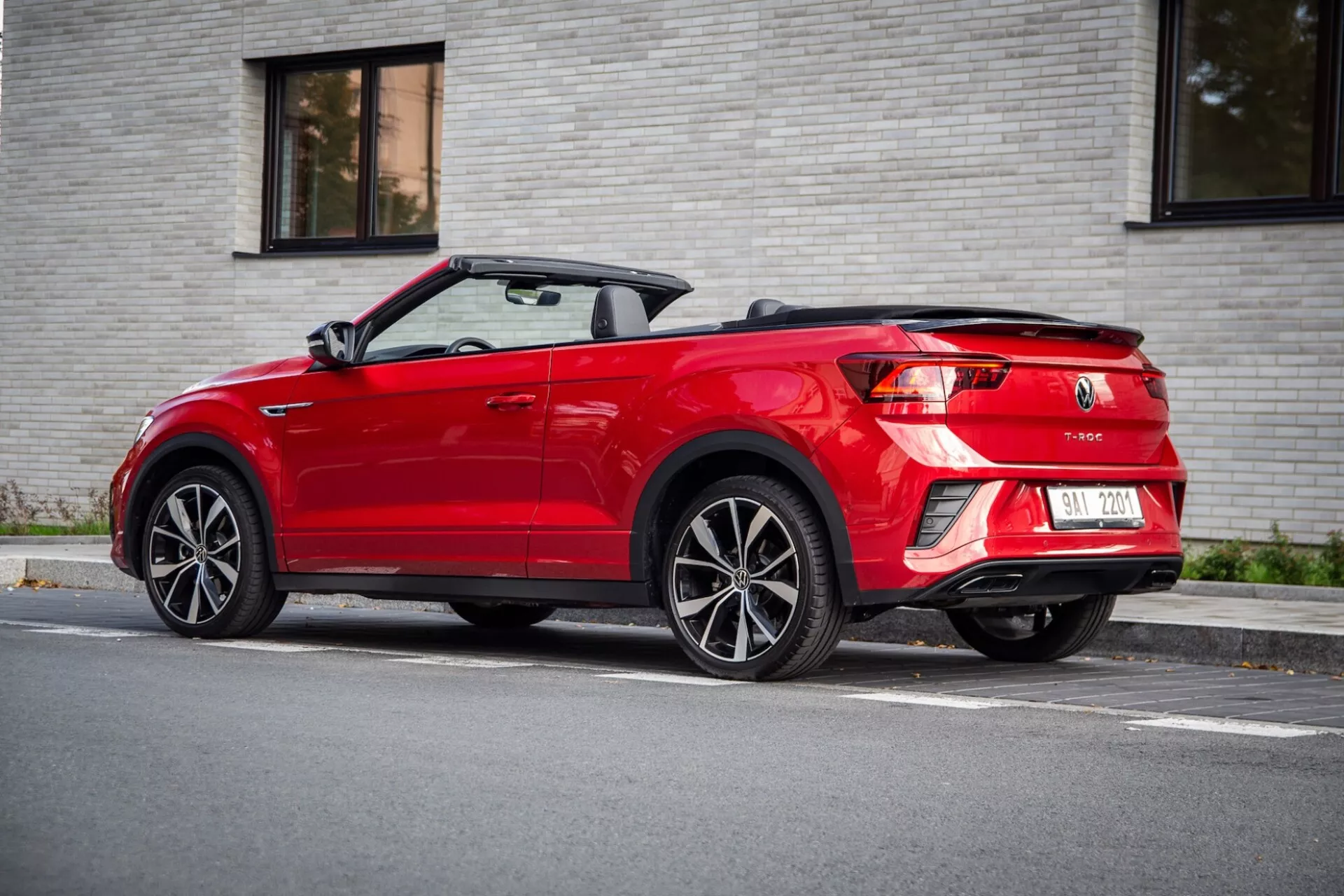 Tříčtvrteční zadní pohled na červený VW T-Roc Cabriolet z roku 2022 se sklopenou měkkou střechou, předvádějící LED zadní světla, dvojité výfuky a kola z lehkých slitin s více paprsky, parkovaný na ulici ve slabém denním světle