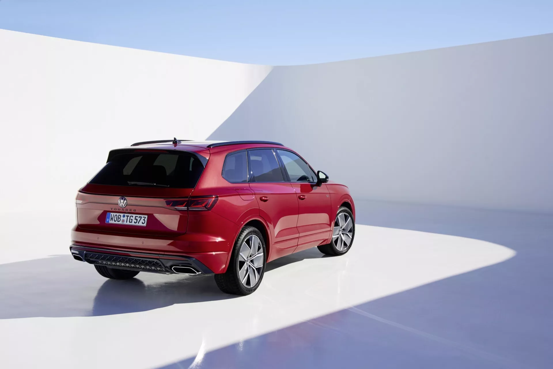 Fotografie modelu VW Touareg III facelift 2023 z pohledu ze zadního tříčtvrtečního úhlu zdůrazňující pravou stranu, ve studiu se spojitým pozadím, předvádějící jeho lesklý červený povrch, aerodynamický design s ostrými liniemi karoserie, LED zadní svítilny, zadní stěrač, spoiler montovaný na střeše a slitinová kola.