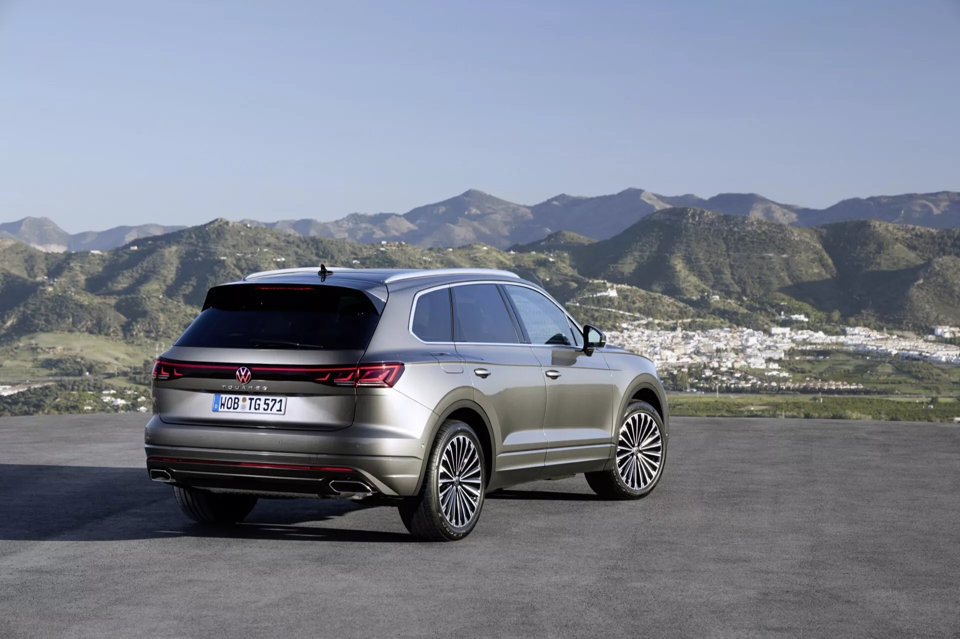 VW Touareg 2023 (III Facelift) SUV zaparkované venku s malebným pozadím kopce a předměstí, zobrazující pravý zadní čtvrtletní a boční profil s důrazem na zadní světla, čáru střechy a linie kontur karoserie. Vlastnosti zahrnují velká slitinová kola, panoramatické střešní okno, zadní LED světla a prémiové prvky stylingu pro jízdu v terénu.
