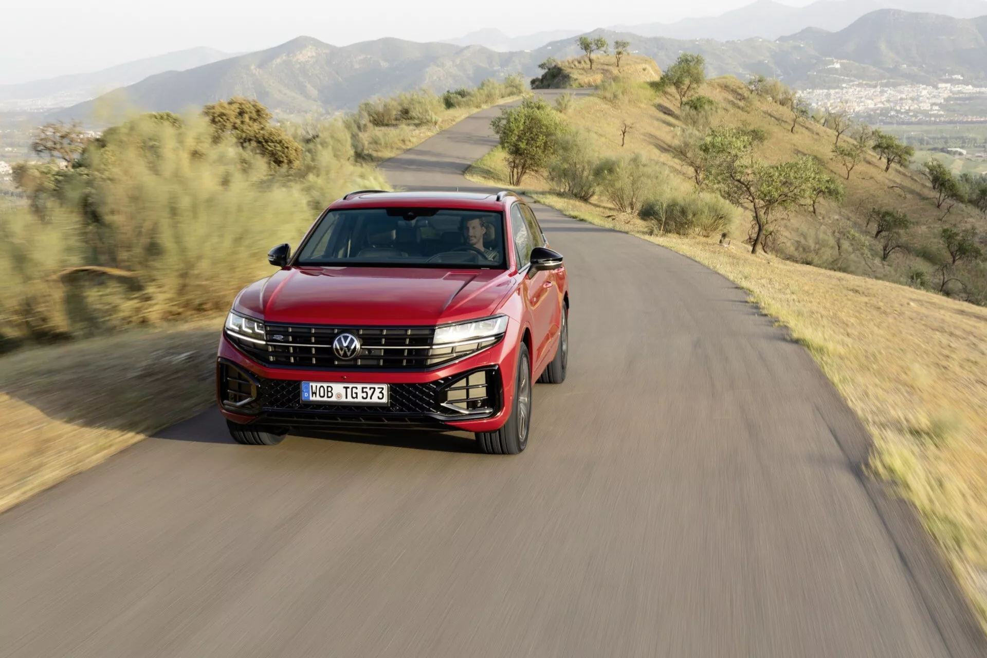 VW Touareg 2023 (III Facelift) crossover/SUV jedoucí po klikaté silnici na kopci, s LED světlomety, slitinovými koly, střešními nosiči, panoramatickou střechou, dvoubarevným lakem a zrcátky se směrovými signály, zachycen z ptačí perspektivy ve dne