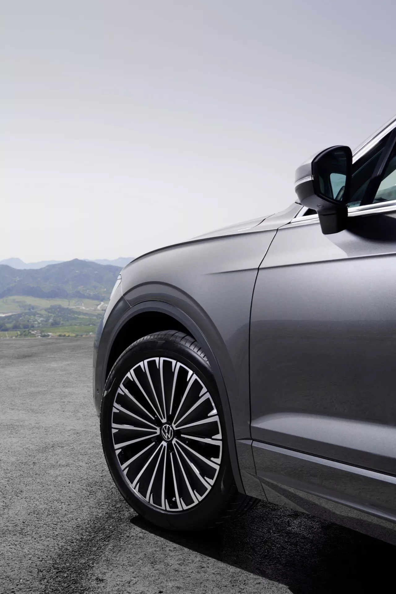 Detailní pohled na přední stranu řidičovy strany Volkswagenu Touareg 2023 (III Facelift) s velkým, stylovým slitinovým kolem s komplexním vícepaprskovým designem, odrážející kvalitu prémiového vozidla na pozadí horské krajiny.