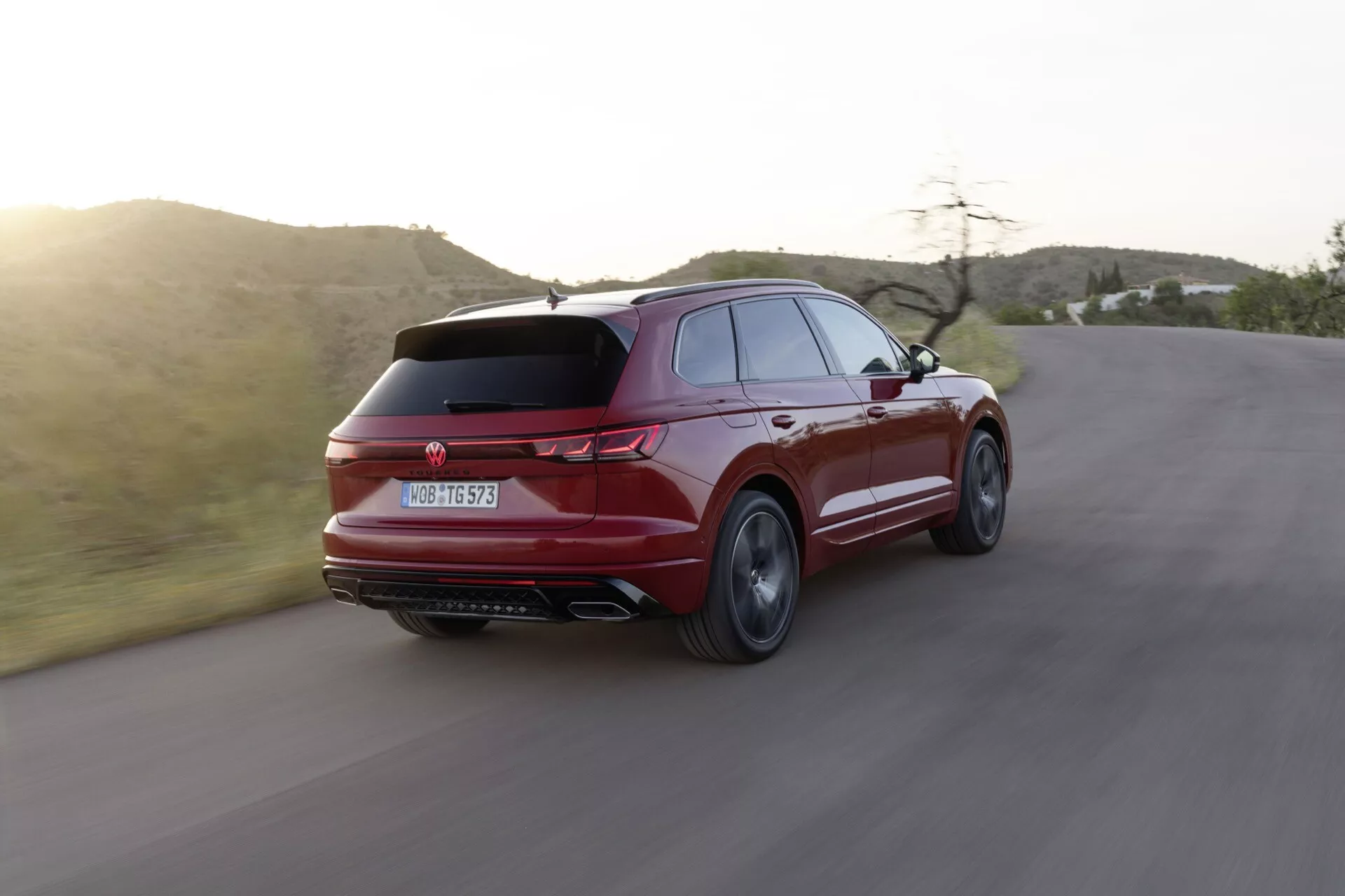 Facelift Volkswagen Touareg III 2023 zachycený v dynamickém tříčtvrtečním zadním pohledu na zakřivené silnici, zdůrazňující jeho sportovně elegantní design, štíhlý zadní spoiler, charakteristická zadní světla, dvojité výfukové trubky a vícepaprsková slitinová kola na pozadí semi-aridní krajiny během zlaté hodiny